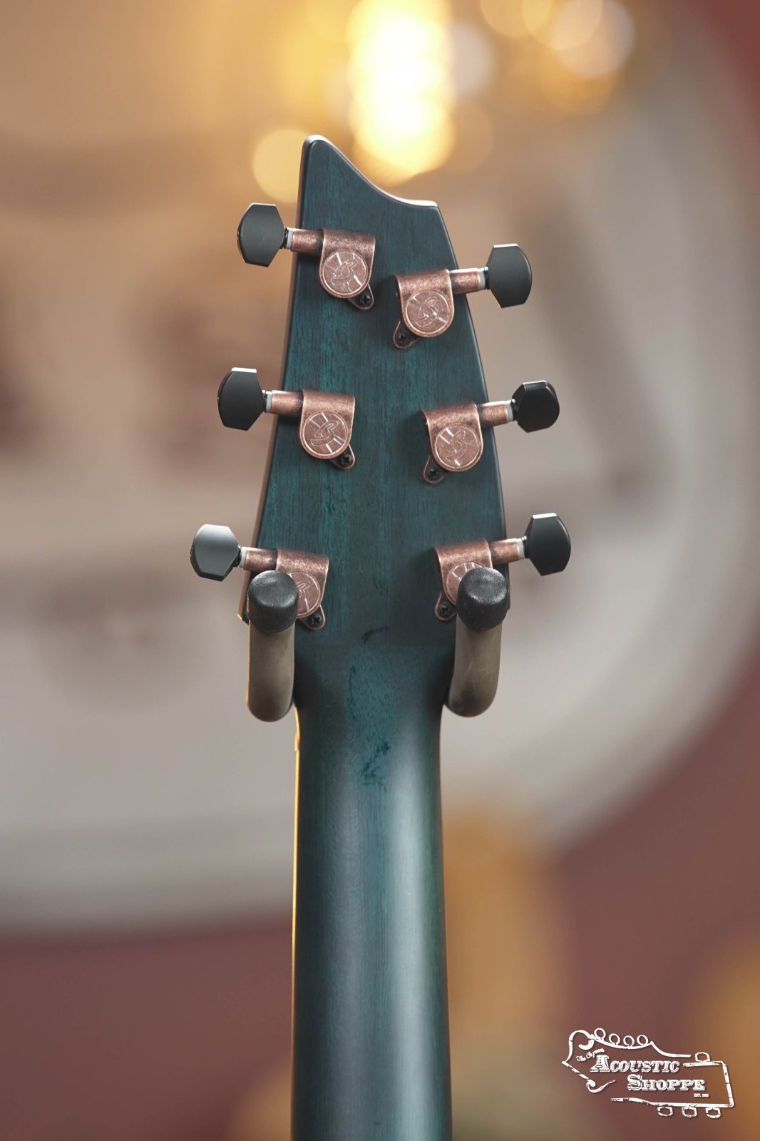 Close-up of the Breedlove Rainforest S Concert Papillon All Mahogany Cutaway Acoustic Guitar headstock with six tuning pegs, blue-green finish, and warm blurred background. Acoustic Shoppe logo is in the bottom right corner.