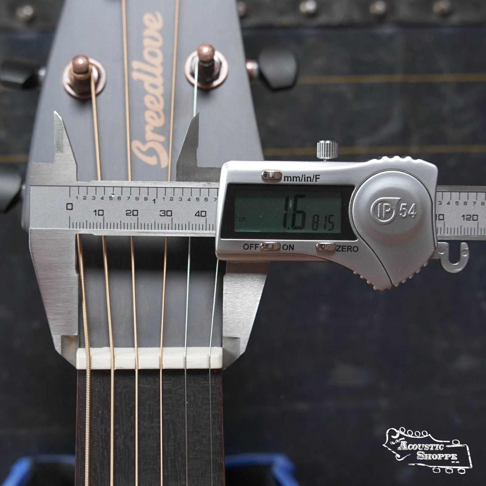 A digital caliper measures the nut width of a Breedlove Rainforest S Concert Papillon All Mahogany Cutaway Acoustic Guitar w/Fishman Pickup, showing 1.6815 inches. The headstock, strings, and “Acoustic Shoppe” logo are also visible.