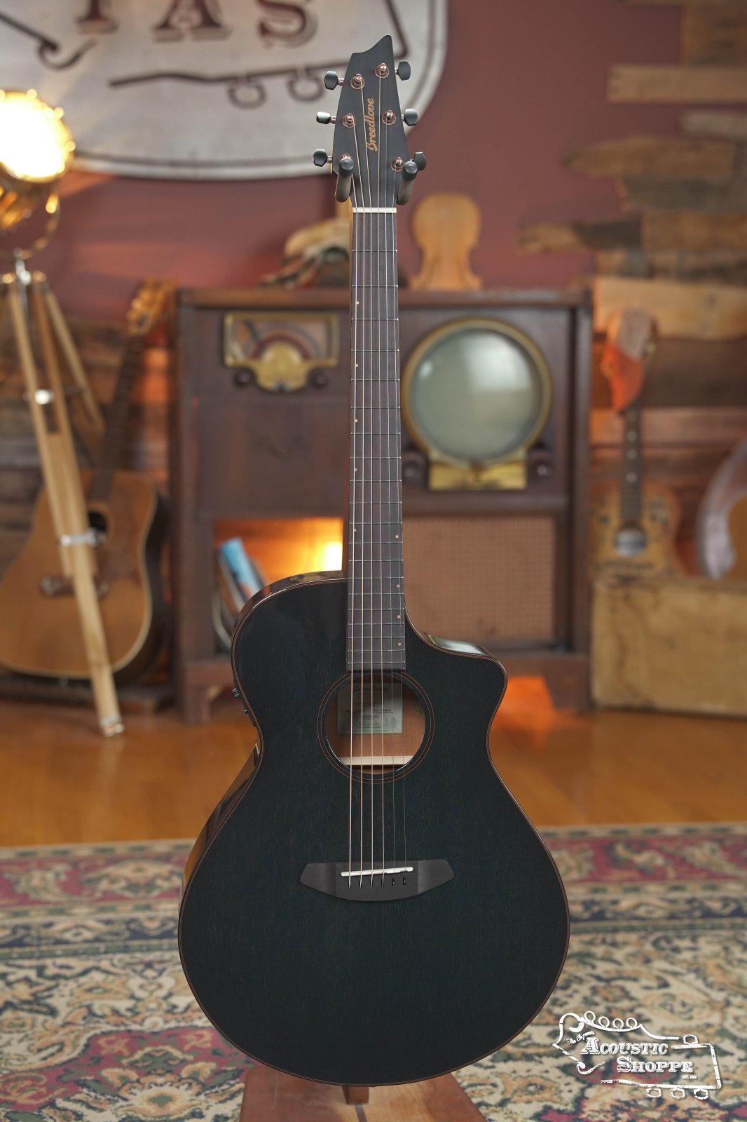 The Breedlove Rainforest S Concert Papillon All Mahogany Cutaway Acoustic Guitar w/Fishman Pickup #6534 stands upright on a patterned rug, its rich African Mahogany exuding warmth in a cozy shop accented by vintage radios and wooden decor.