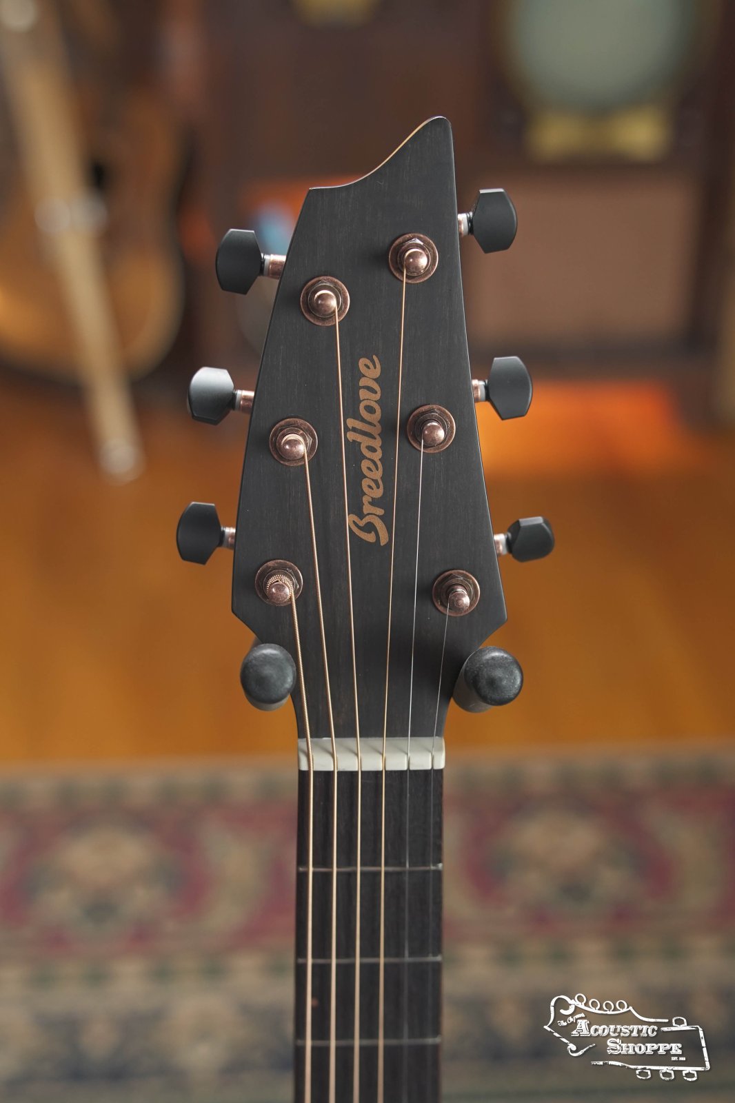 Close-up of a Breedlove Rainforest S Concert Papillon All Mahogany Cutaway Acoustic Guitar w/Fishman Pickup headstock with six tuning pegs and bronze hardware. The script logo, blurred wood background, and EcoTonewood craftsmanship are shown.
