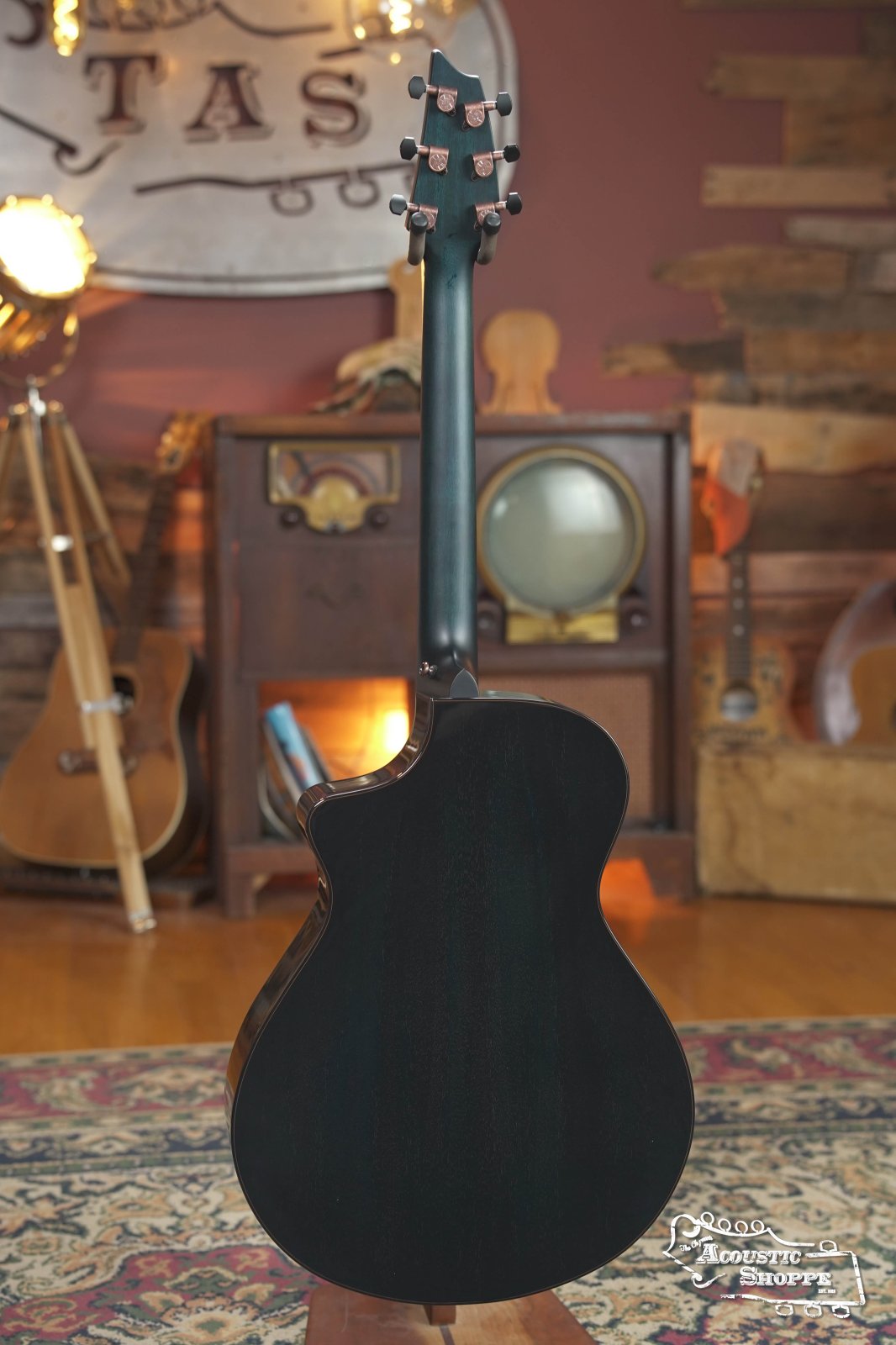 A Breedlove Rainforest S Concert Papillon All Mahogany Cutaway Acoustic Guitar w/Fishman Pickup is displayed from the back on a patterned rug in a warmly lit, vintage-themed room with an old radio, TV, and wooden decor in the background.