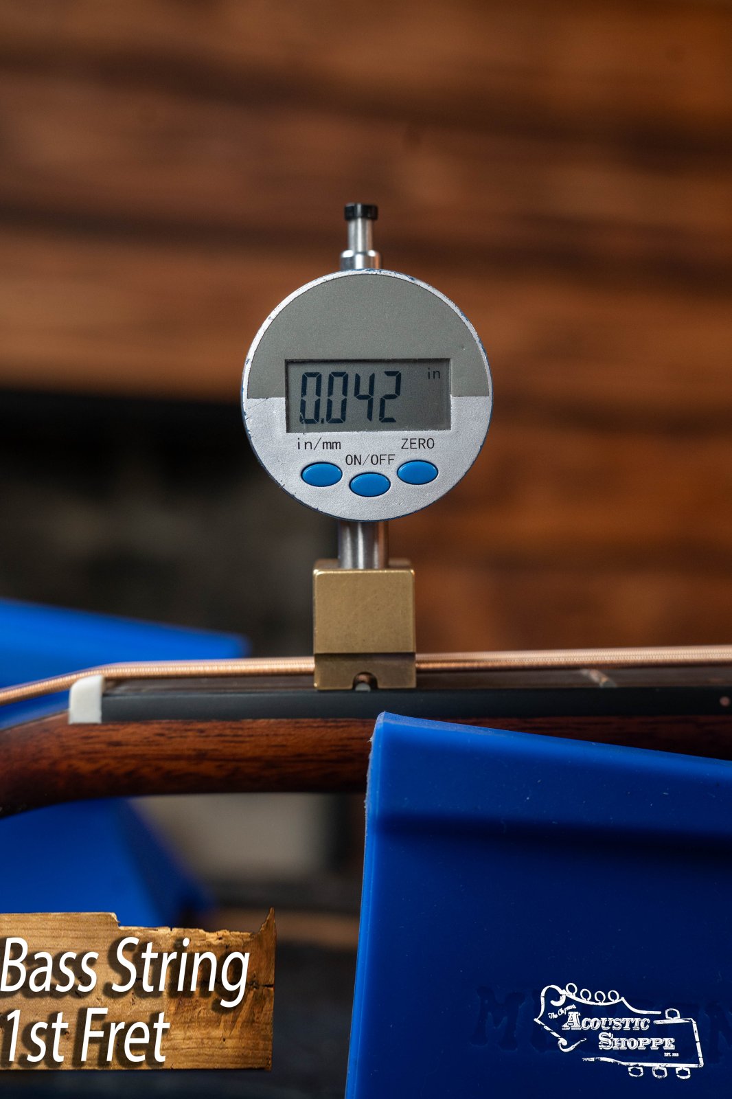 A digital gauge shows 0.042 inches at the 1st fret of a Breedlove Organic Solo Pro Concerto Red Cedar/African Mahogany Acoustic Bass, with “Bass String 1st Fret” and “Acoustic Shoppe” over a blurred wood background—ideal for setup checks.