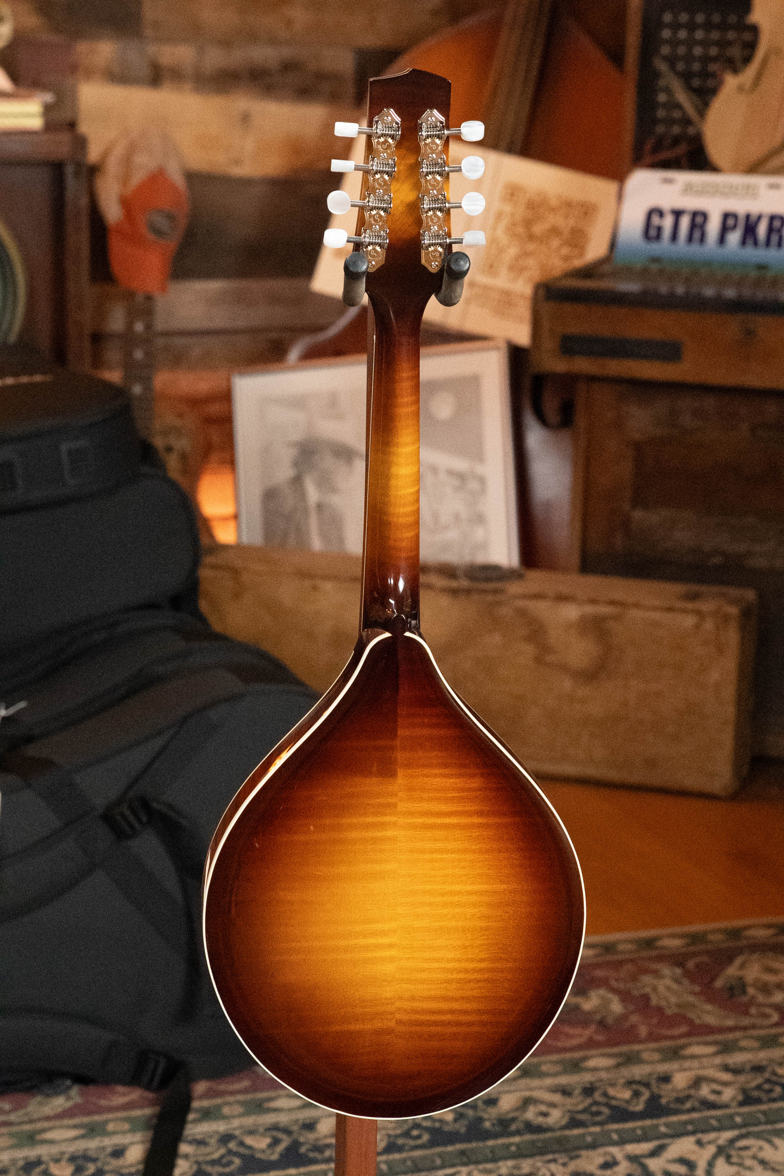 The Bulas Carpathian Spruce/Curly Maple Cremona Sunburst A-Style Mandolin (#8129) is displayed from the back, showing its glossy wood-grain body and headstock with tuning pegs in a room with musical decor and wooden furniture.