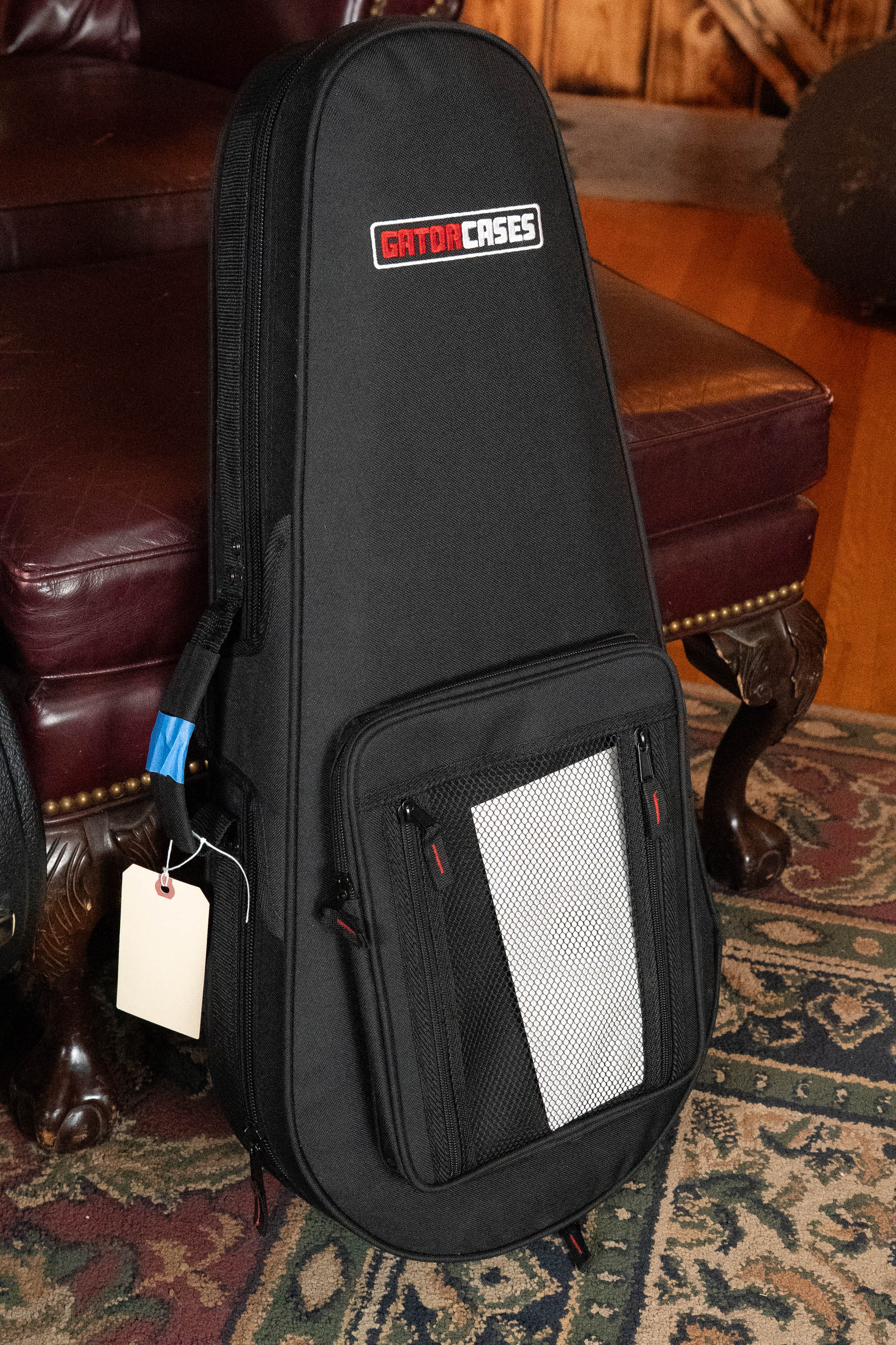 A black Gator Cases gig bag with front pockets and a white mesh section stands upright on a rug next to a red leather chair, ready to protect your Bulas Carpathian Spruce/Curly Maple Cremona Sunburst A-Style Mandolin #8129.