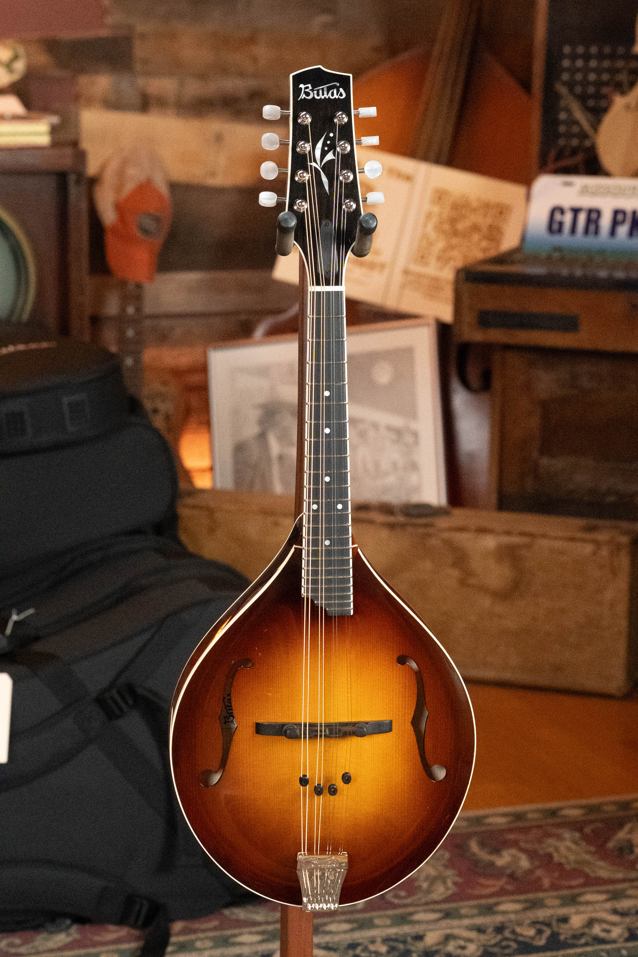 A Bulas Carpathian Spruce/Curly Maple Cremona Sunburst A-Style Mandolin #8129 is displayed on a stand in a cozy, rustic room with wood furnishings, music gear, and decor in the background.