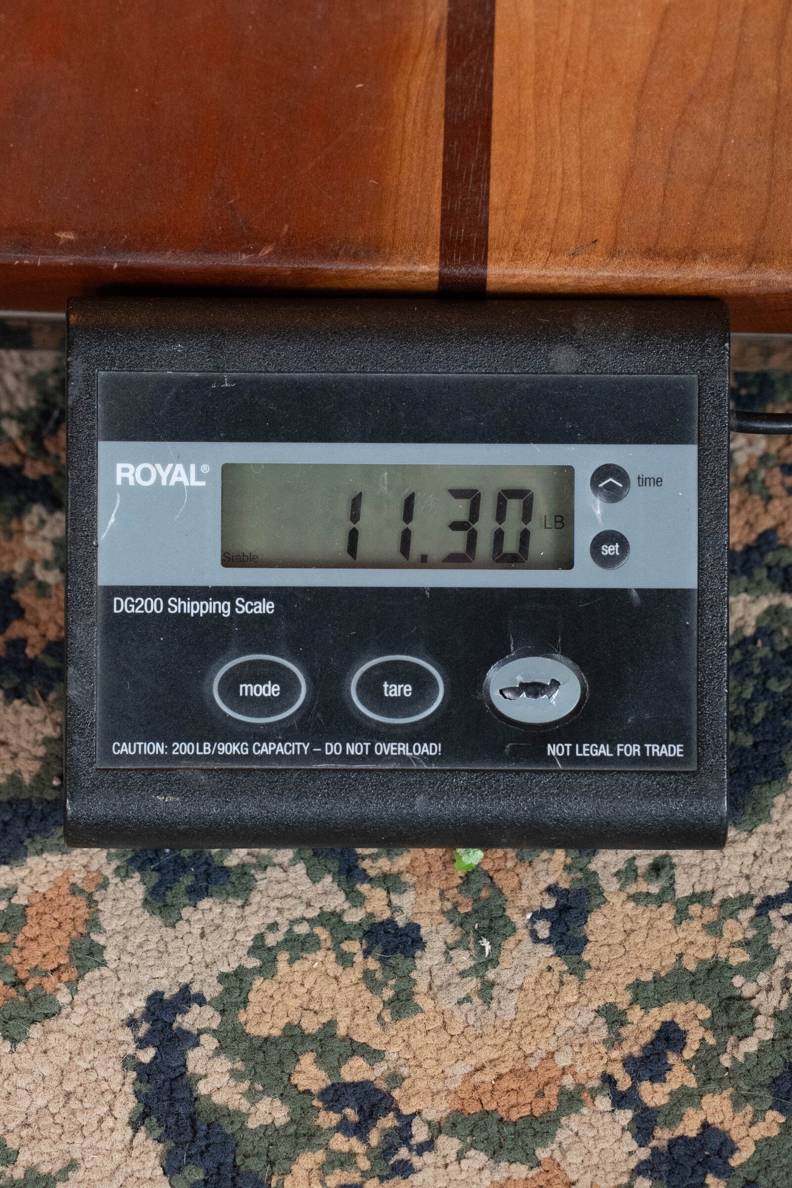 A Bulas Standard 5-String Tobacco Sunburst Resonator Banjo #0676 rests partly over a digital shipping scale on patterned carpet. The scale, featuring mode, tare, and set buttons, displays a weight of 11.30 pounds.