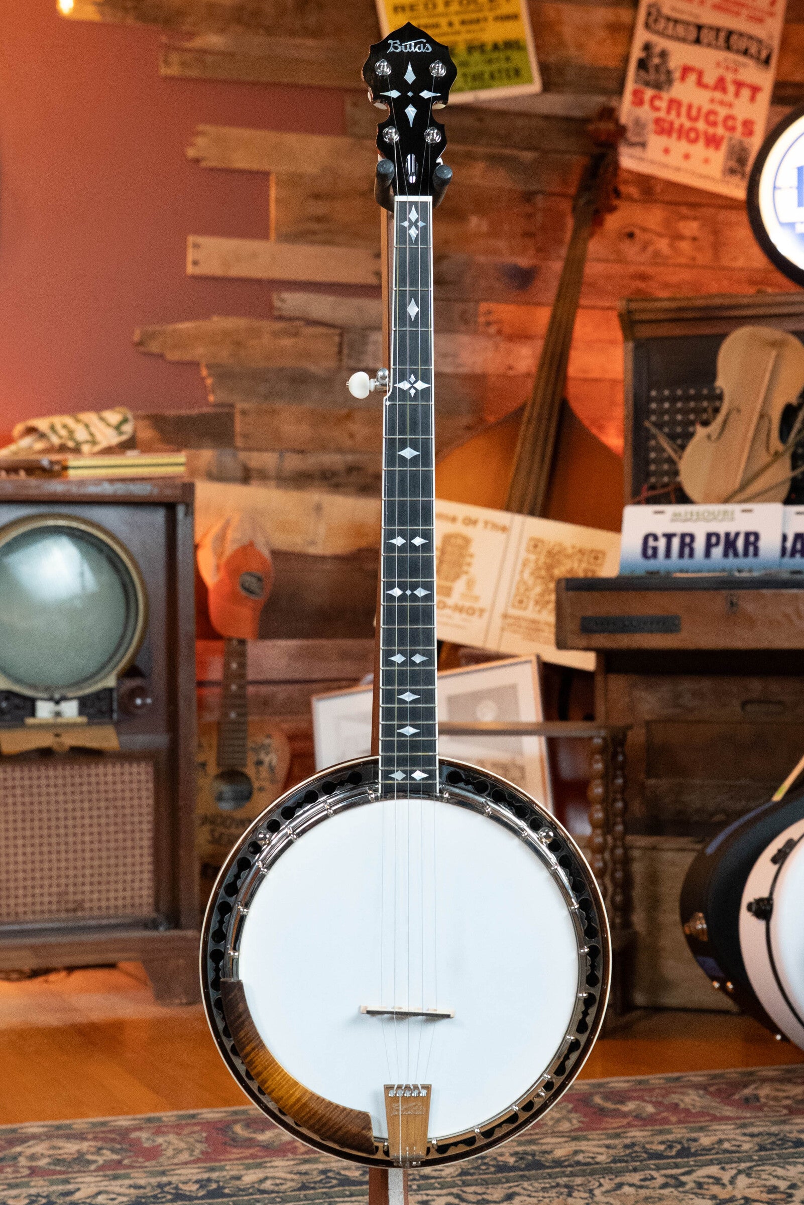 The Bulas Standard 5-String Tobacco Sunburst Resonator Banjo #0676 by Bulas stands upright in a rustic room among vintage musical instruments, an old TV, and retro music posters.