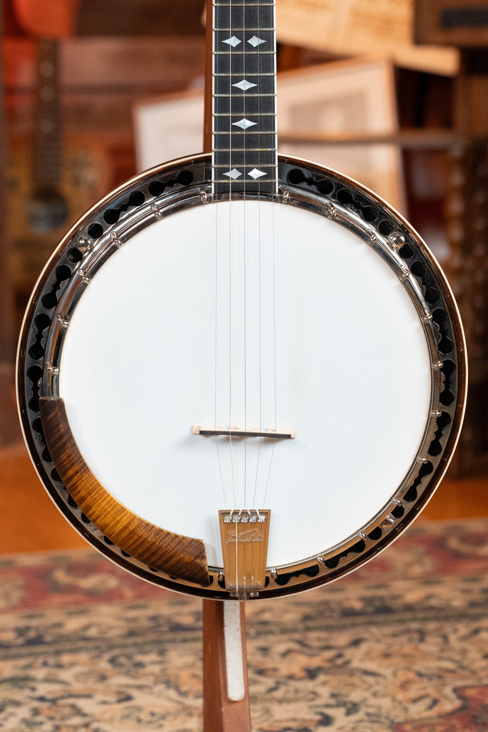 A close-up of the Bulas Standard 5-String Tobacco Sunburst Resonator Banjo #0676 by Bulas shows its round white drum head, strings, black fretboard with white inlays, and a maple rim with bronze tone ring on a stand.