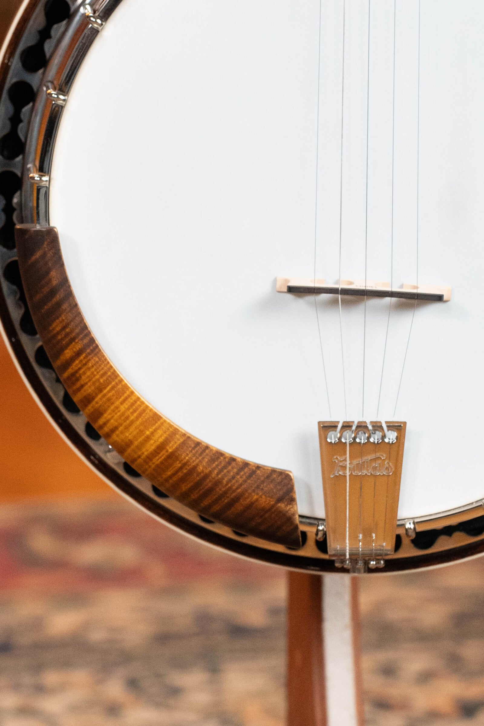 Close-up of the Bulas Standard 5-String Tobacco Sunburst Resonator Banjo #0676 by Bulas, highlighting its white drumhead, wooden rim, metal bridge and tailpiece; strings extend upward against a blurred background.