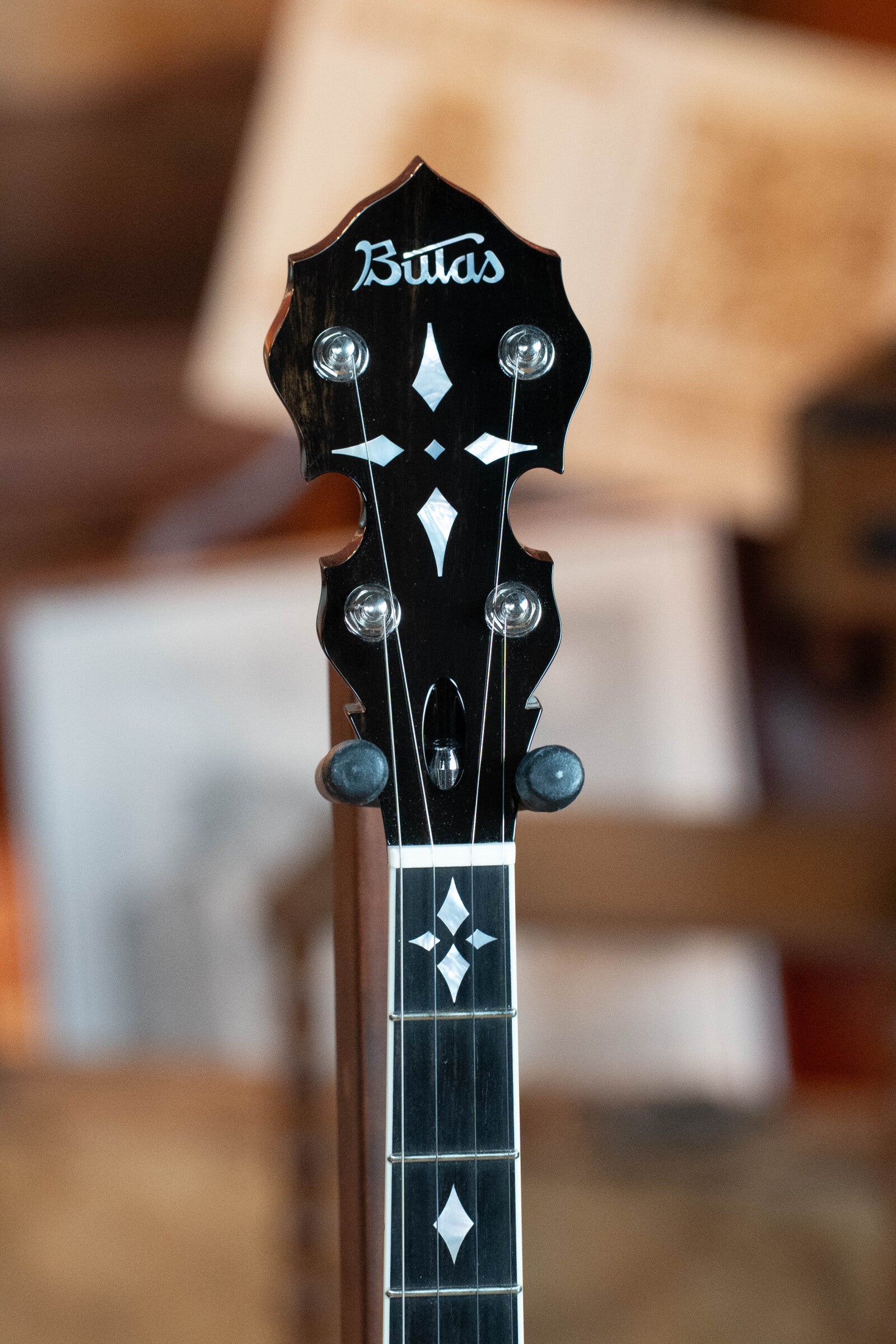 Close-up of the headstock and upper neck of a Bulas Standard 5-String Tobacco Sunburst Resonator Banjo #0676, showing decorative inlays and a curly maple neck by Bulas, set against a blurred background.