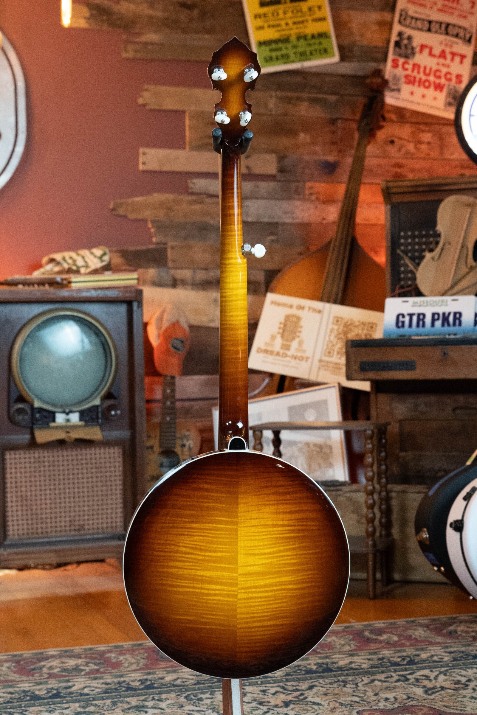The Bulas Standard 5-String Tobacco Sunburst Resonator Banjo #0676 stands upright in a warmly lit room filled with vintage instruments, posters, and an old TV set, its curly maple neck contrasting beautifully against the wooden wall.