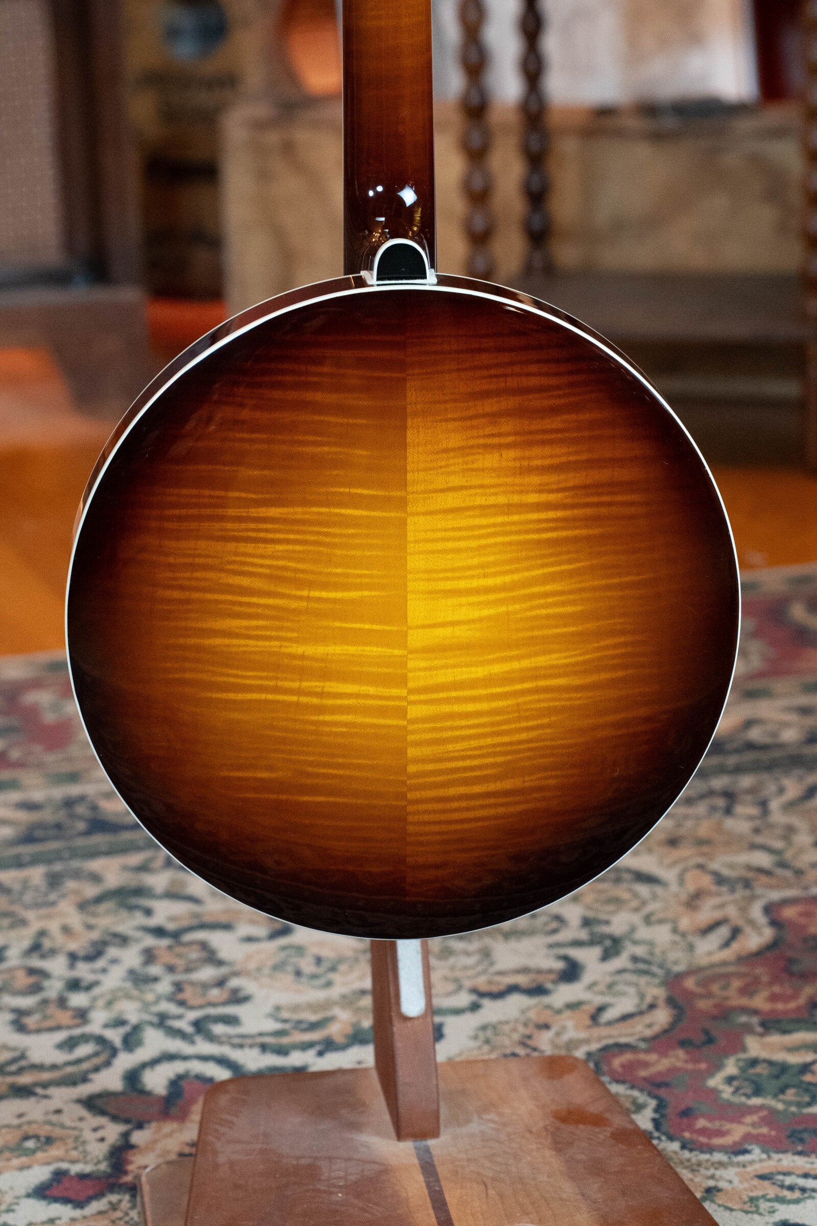 A close-up of the Bulas Standard 5-String Tobacco Sunburst Resonator Banjo #0676 by Bulas, showcasing its glossy sunburst finish as it rests on a wooden stand over a patterned rug.