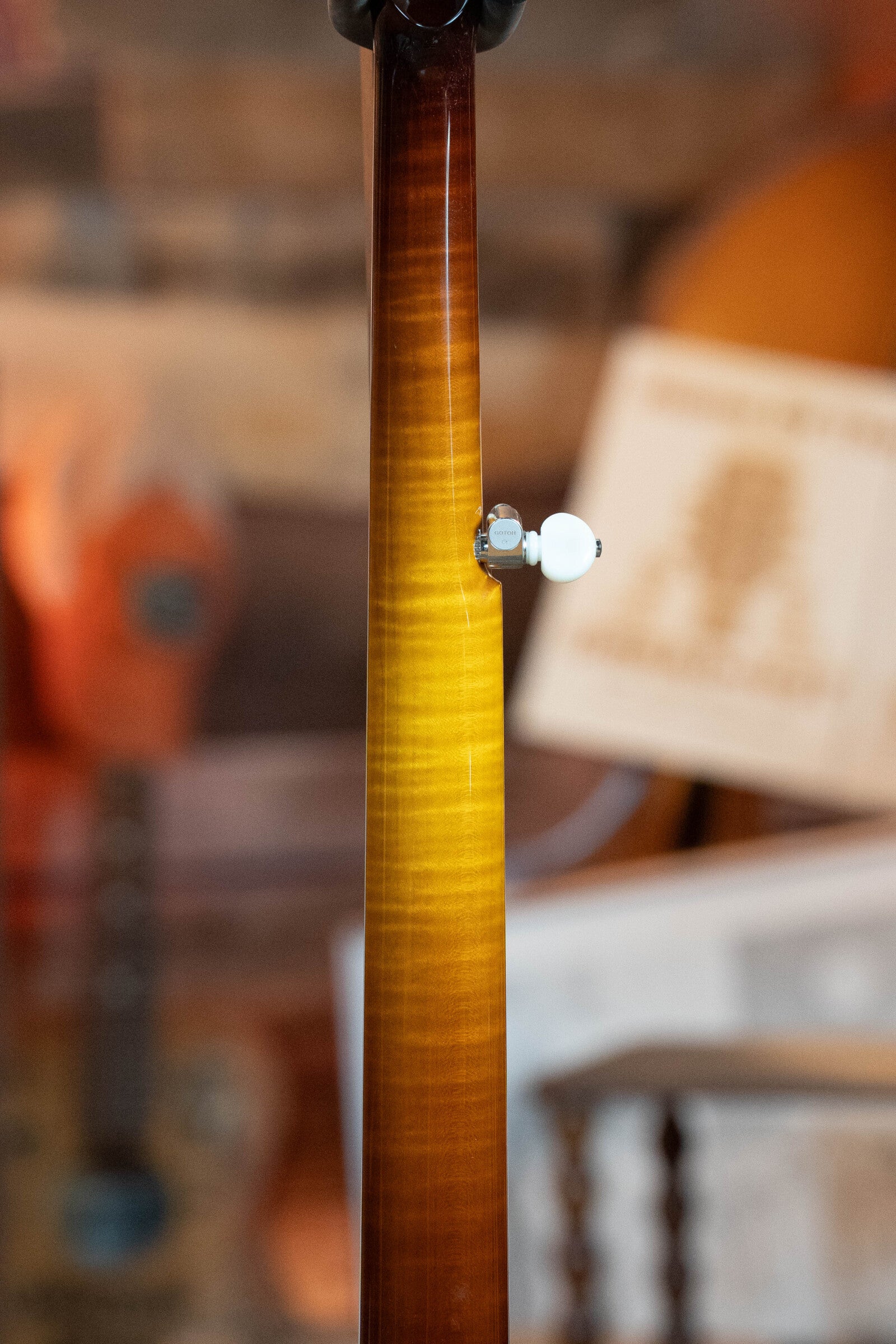 A close-up of the Bulas Standard 5-String Tobacco Sunburst Resonator Banjo #0676’s curly maple neck highlights a white tuning peg, with other wooden instruments and sheet music blurred in the background.