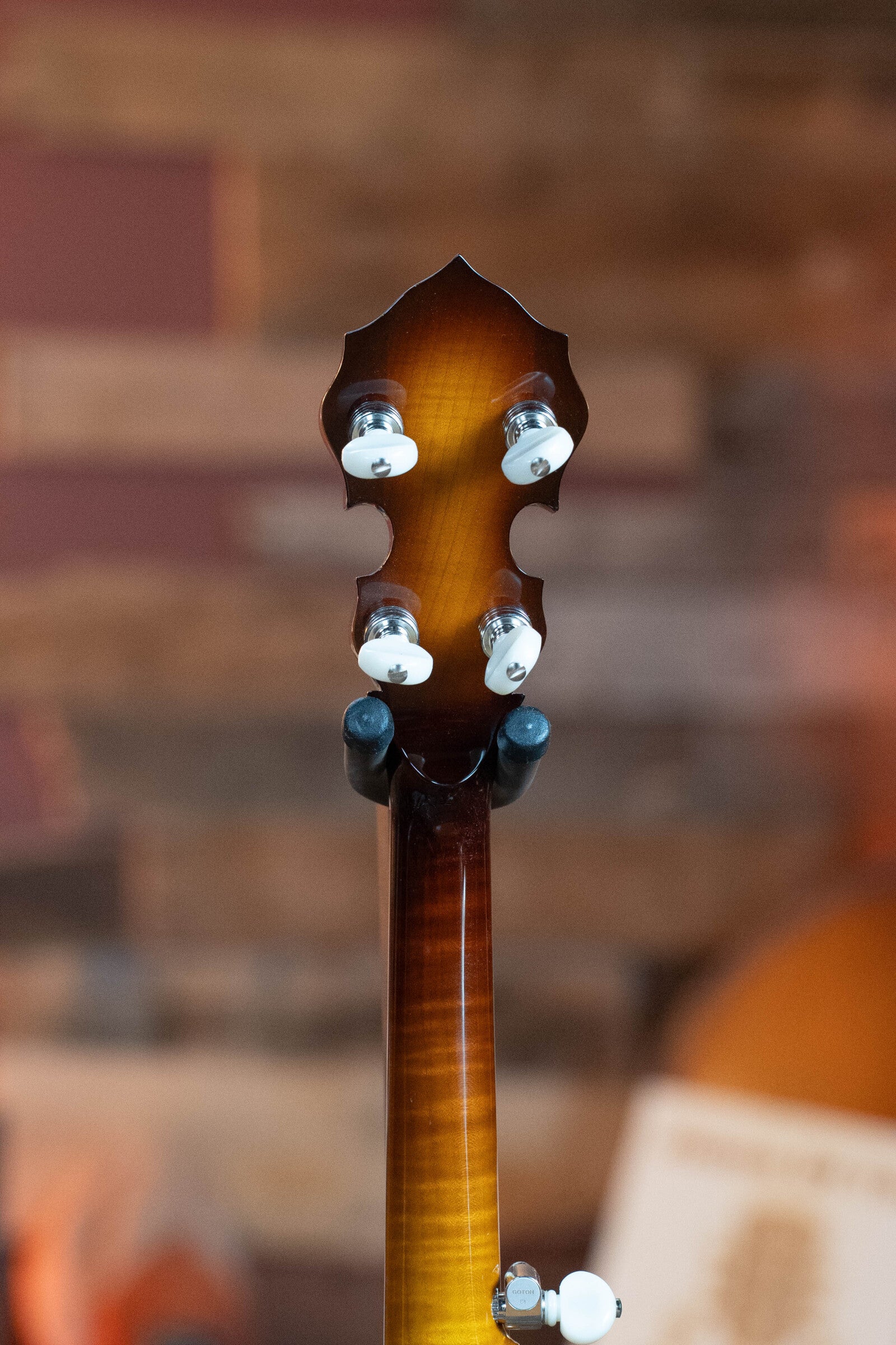 Close-up of the back of a Bulas Standard 5-String Tobacco Sunburst Resonator Banjo #0676 headstock, showing four silver and white tuning pegs. The blurred background features warm wood tones and soft shapes.