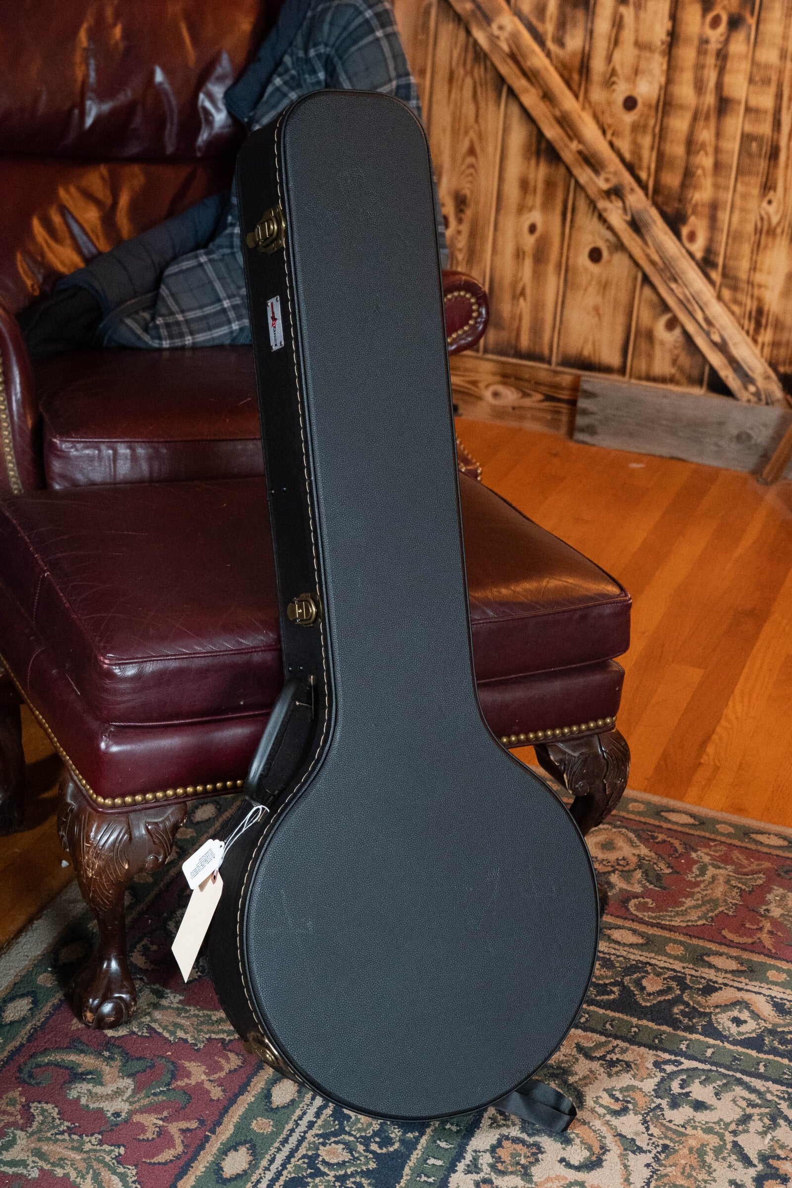 A Bulas Standard 5-String Tobacco Sunburst Resonator Banjo #0676 in its black hard-shell case with handle sits on an ornate red leather chair atop a patterned rug, with wood-paneled walls in the background.