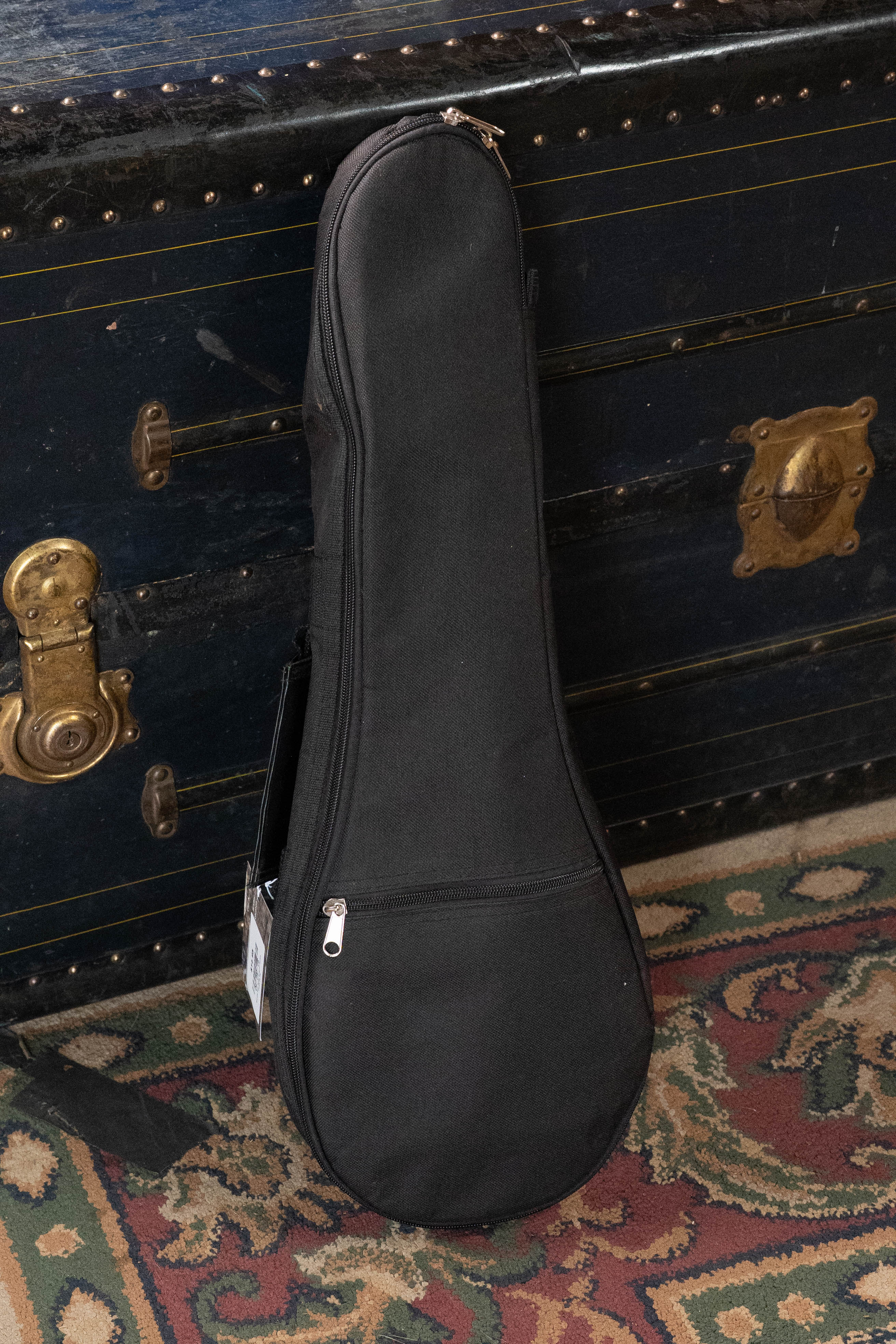 The Guardian 6mm Padded Duraguard Soprano Ukulele Gig Bag, in black with backpack straps and a zipper pocket, stands upright before a vintage trunk atop a patterned rug of green, red, and cream tones.
