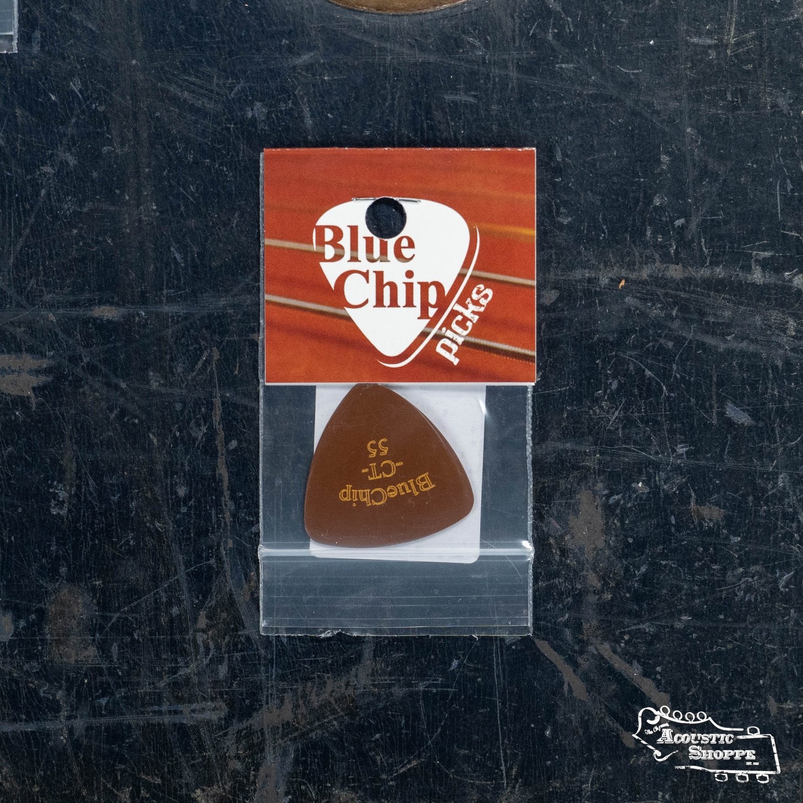 A BlueChip Picks (CT55) guitar pick is labeled Tortoise 55, sealed in clear plastic with a red and white BlueChip label, and rests on a dark, scratched surface with the Folk Acoustic Shoppe logo in the corner.