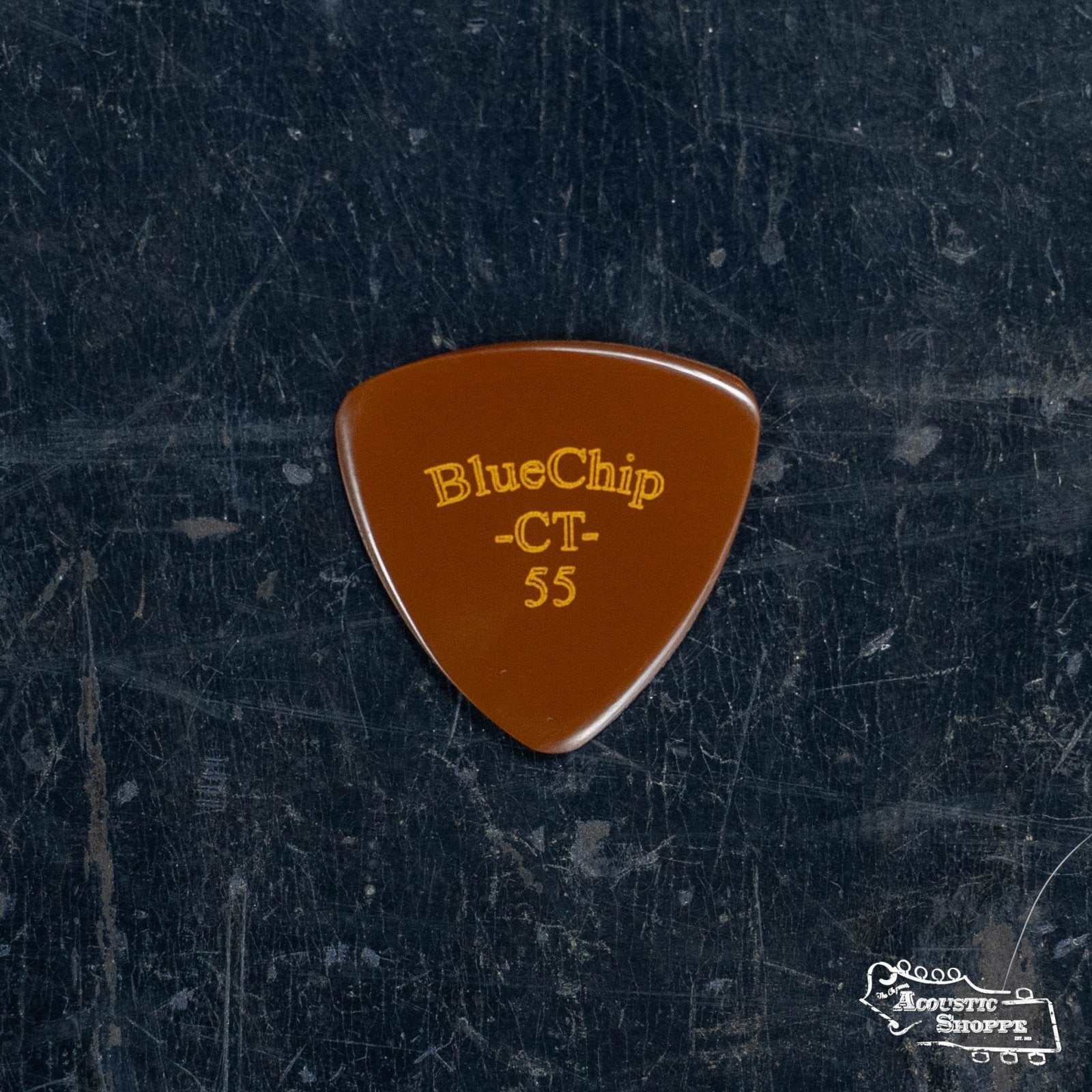 A brown BlueChip Picks (CT55) guitar pick with yellow lettering, made by BlueChip, rests on a worn, dark textured surface. The triangular pick with rounded edges features “BlueChip CT-55,” like the Chris Thile model.