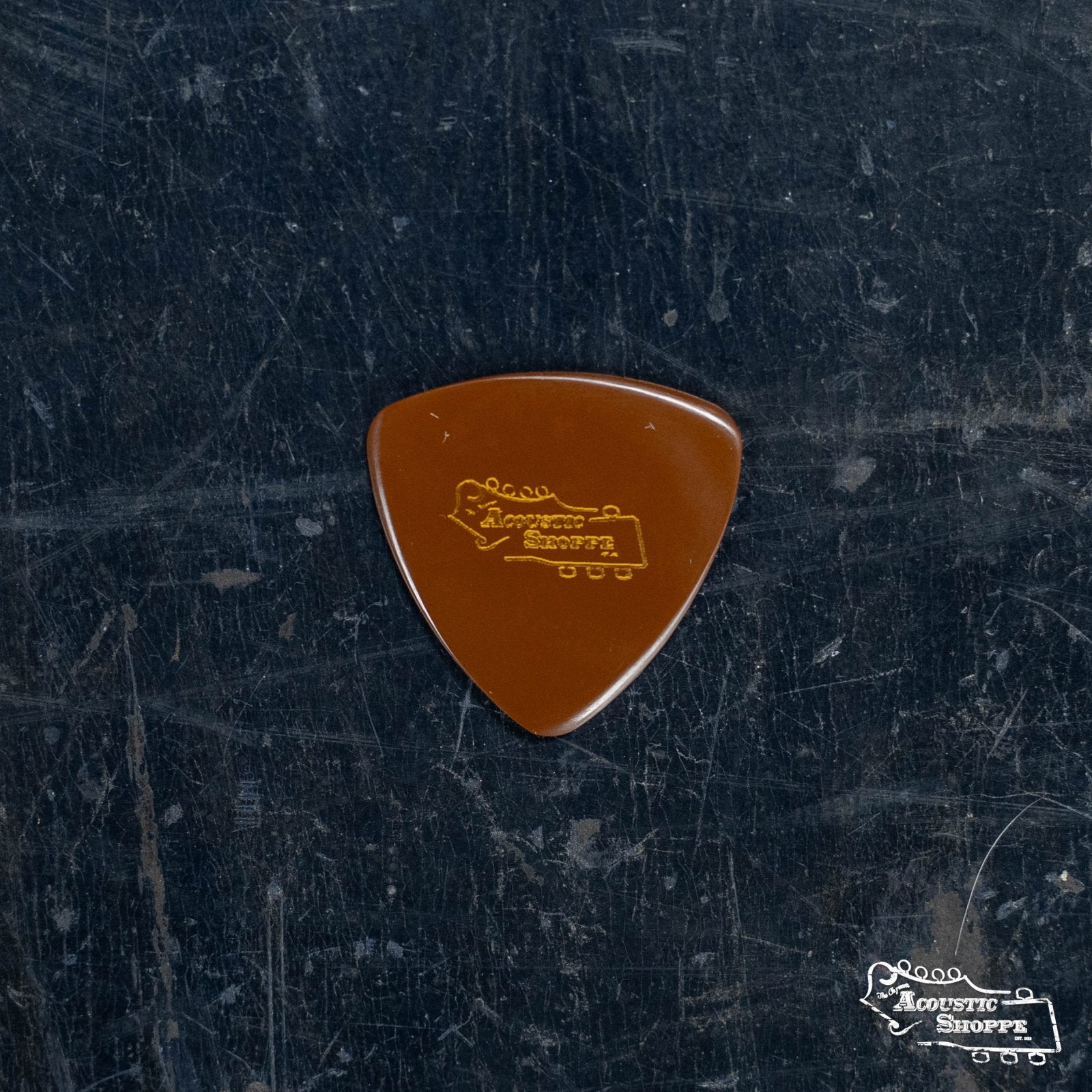 A brown custom BlueChip Picks (CT55) guitar pick by BlueChip, featuring the Acoustic Shoppe and a guitar headstock logo in yellow, resting on a dark textured surfaceāideal for fans of Chris Thileās favorite pick styles.