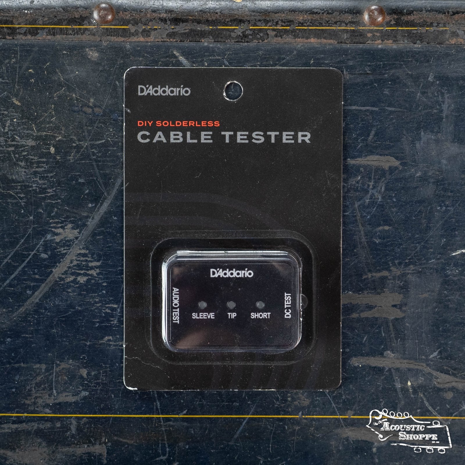 The DAddario Power and Instrument Cable Tester PW-DIYCT-01 is shown in its compact, pocket-sized packaging—black with white and red text—displayed against a worn, dark blue metal surface. The tester is visible through a clear plastic window.