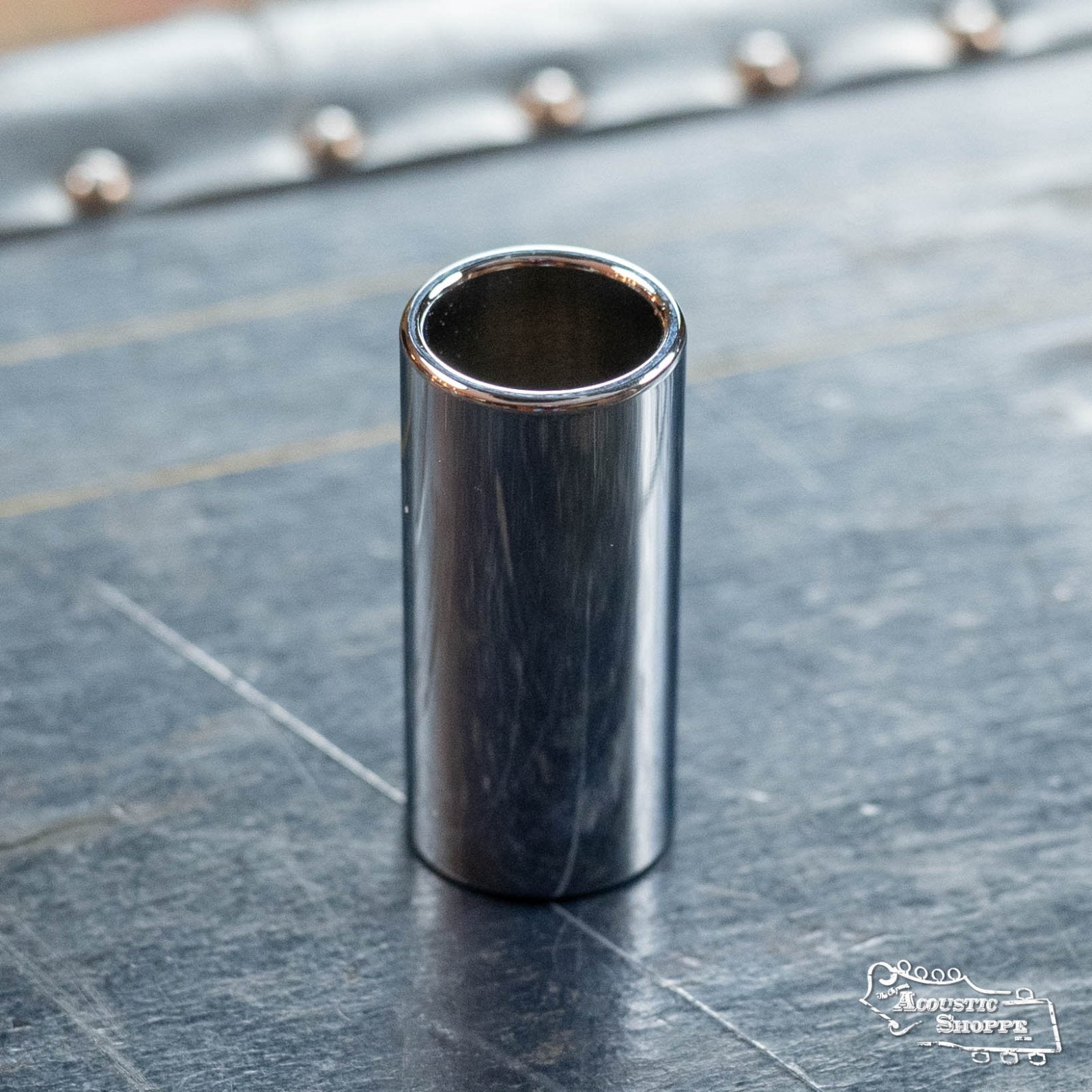 A DAddario Chrome-Plated Brass Guitar Slide Large (PWCBS-SL) stands upright on a dark wooden surface, gleaming like polished chrome. The Acoustic Shoppe logo appears in the bottom right corner.
