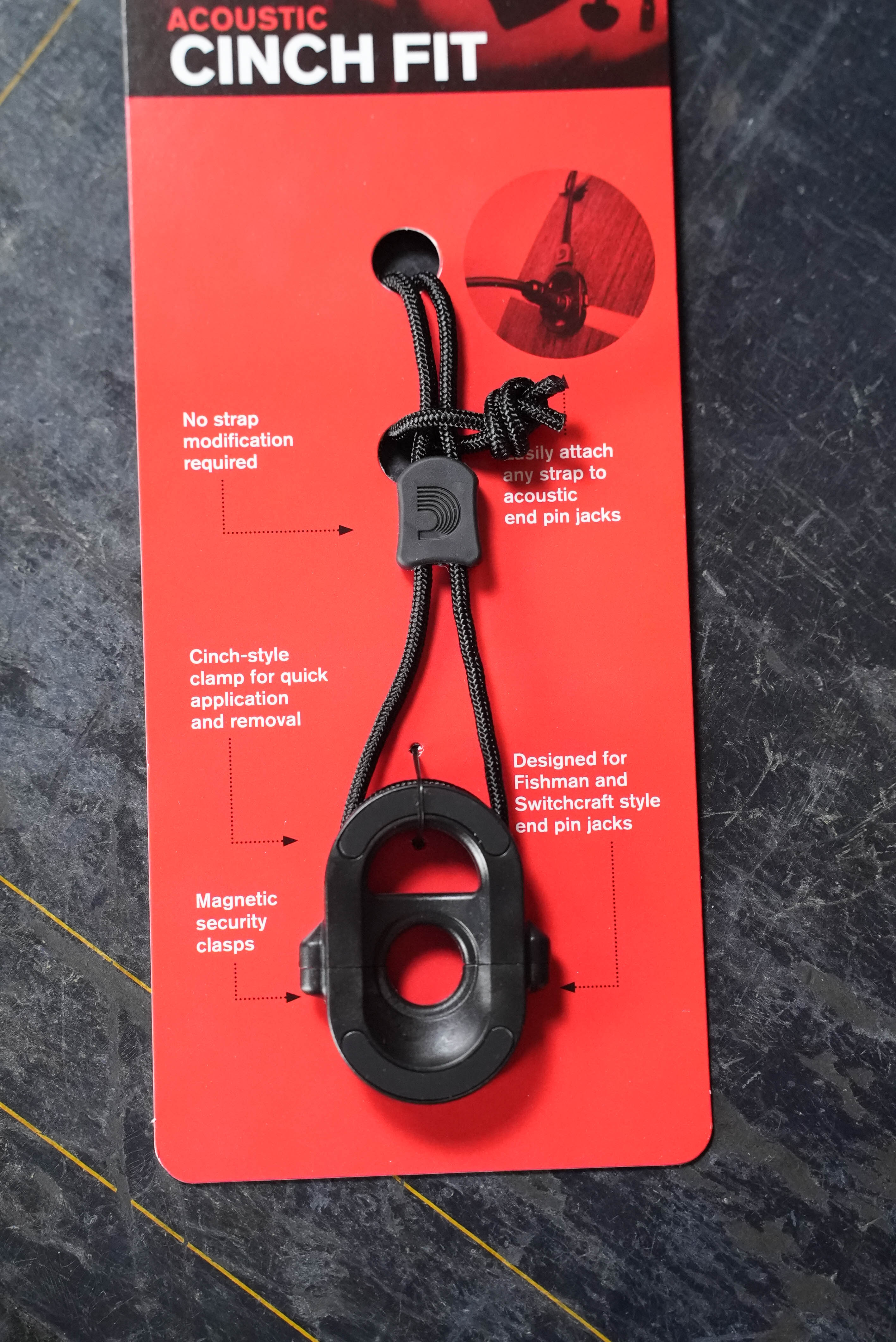 A red card features the D'Addario PW-AJL-01 CinchFit: Acoustic Jack Lock, a black, adjustable acoustic guitar strap adapter with quick clamp and magnetic clasps—fits endpin jack setups, requires no strap modification.