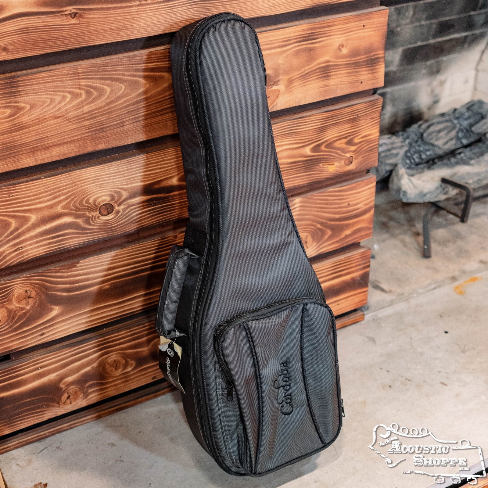A Cordoba Deluxe Gig Bag for Tenor Ukulele/Guilele, made of durable 600D polyester, with a zippered pocket and handle, leans against a wooden wall. Part of a fireplace and concrete floor are visible in the background.