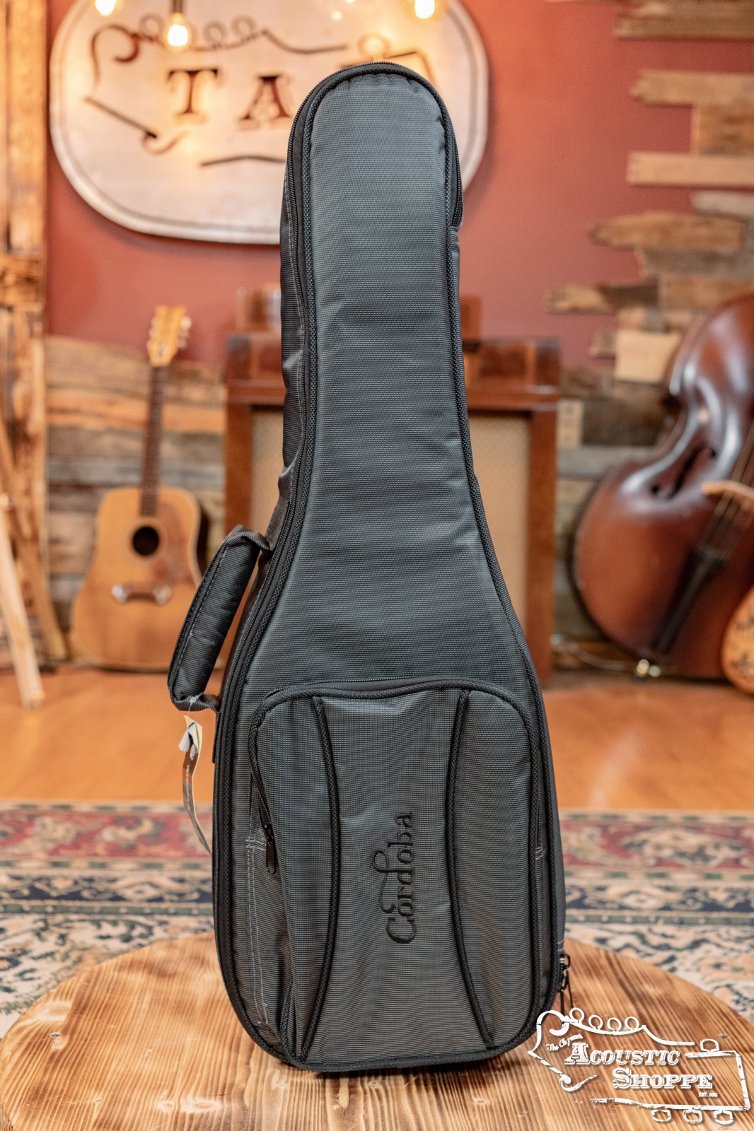 A Cordoba Deluxe Gig Bag for Tenor Ukulele/Guilele stands upright on a rug in a cozy music shop, surrounded by wooden instruments like guitars and a double bass, with rustic decor and acoustic accessories in the background.