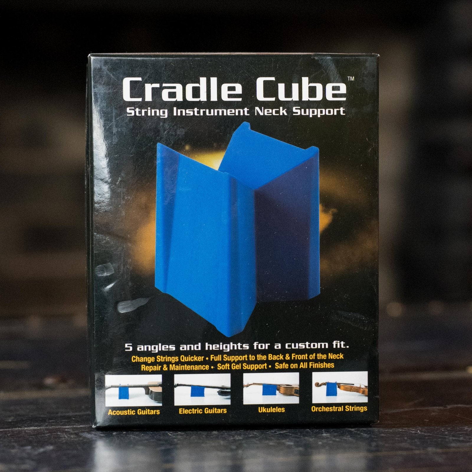 A box labeled Music Nomad Cradle Cube - String Instrument Neck Support (MN206) is shown. The packaging features a blue device by Music Nomad for supporting acoustic guitars, electric guitars, ukuleles, and orchestral string instruments.