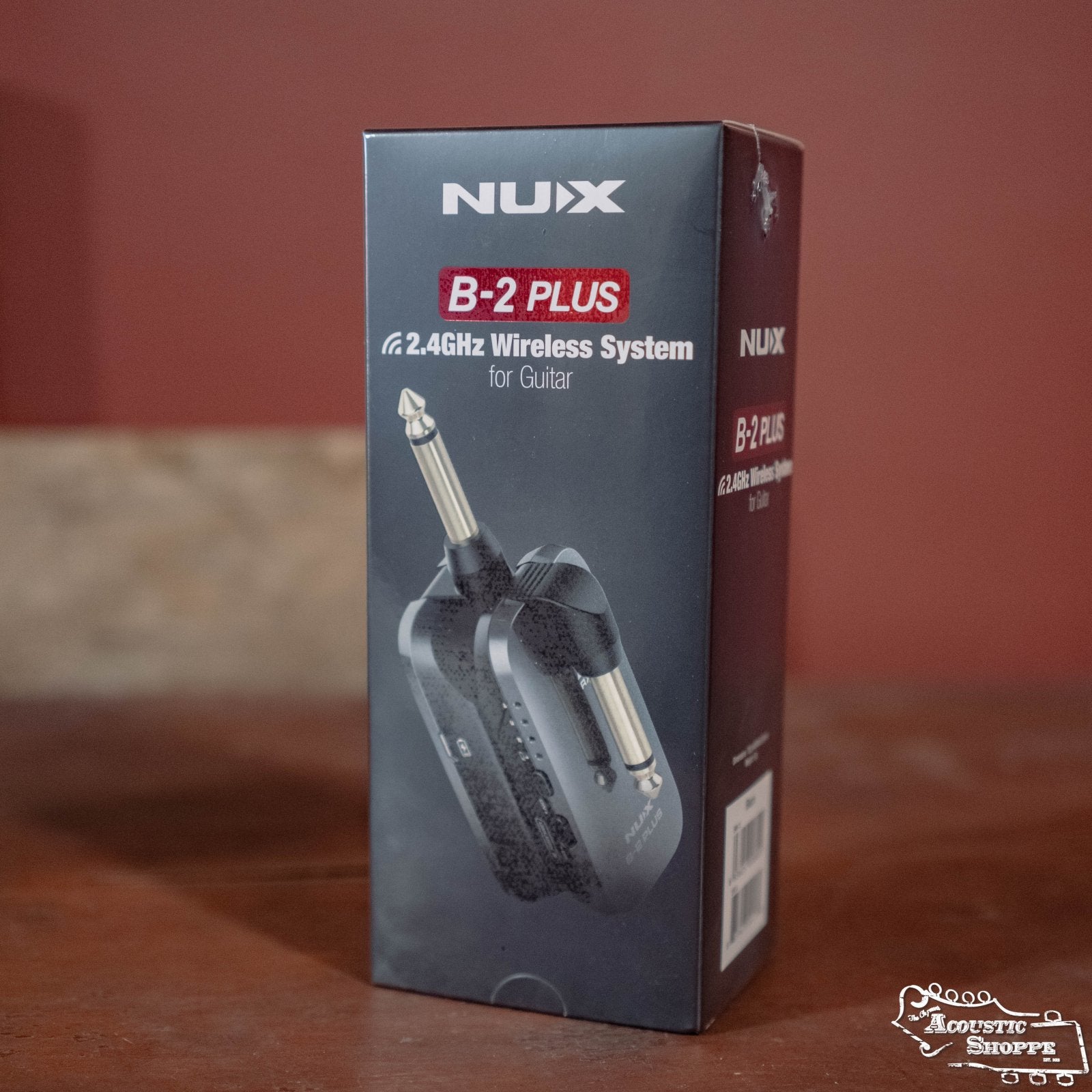 The NUX B-2 Plus Wireless Guitar System for Acoustic or Electric (Black) is displayed in its stylish black and red packaging on a wooden surface, with a blurred brown background highlighting the product.