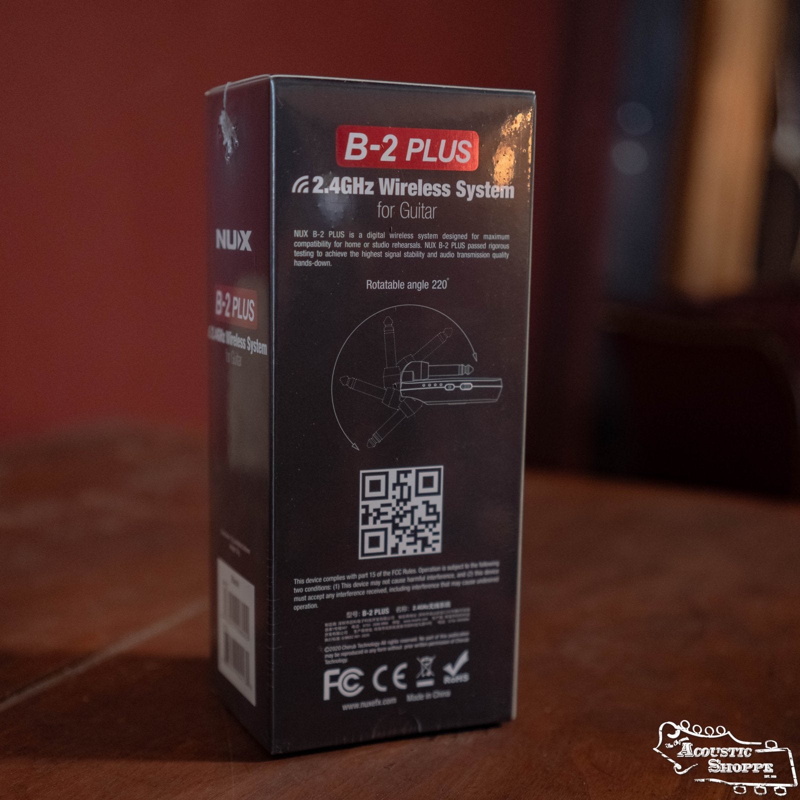 A box of the NUX B-2 Plus Wireless Guitar System for Acoustic or Electric (Black) by NUX is displayed on a wooden table, featuring product details, a diagram, and a QR code on its 2.4GHz wireless packaging.
