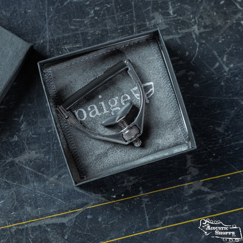 A Paige Musical Products RetroPro 6-String Capo (PP-6-Retro) in black rests in an open, gray-lined box labeled “paige,” alongside two yellow guitar strings and a small Acoustic Shoppe logo on a dark surface.