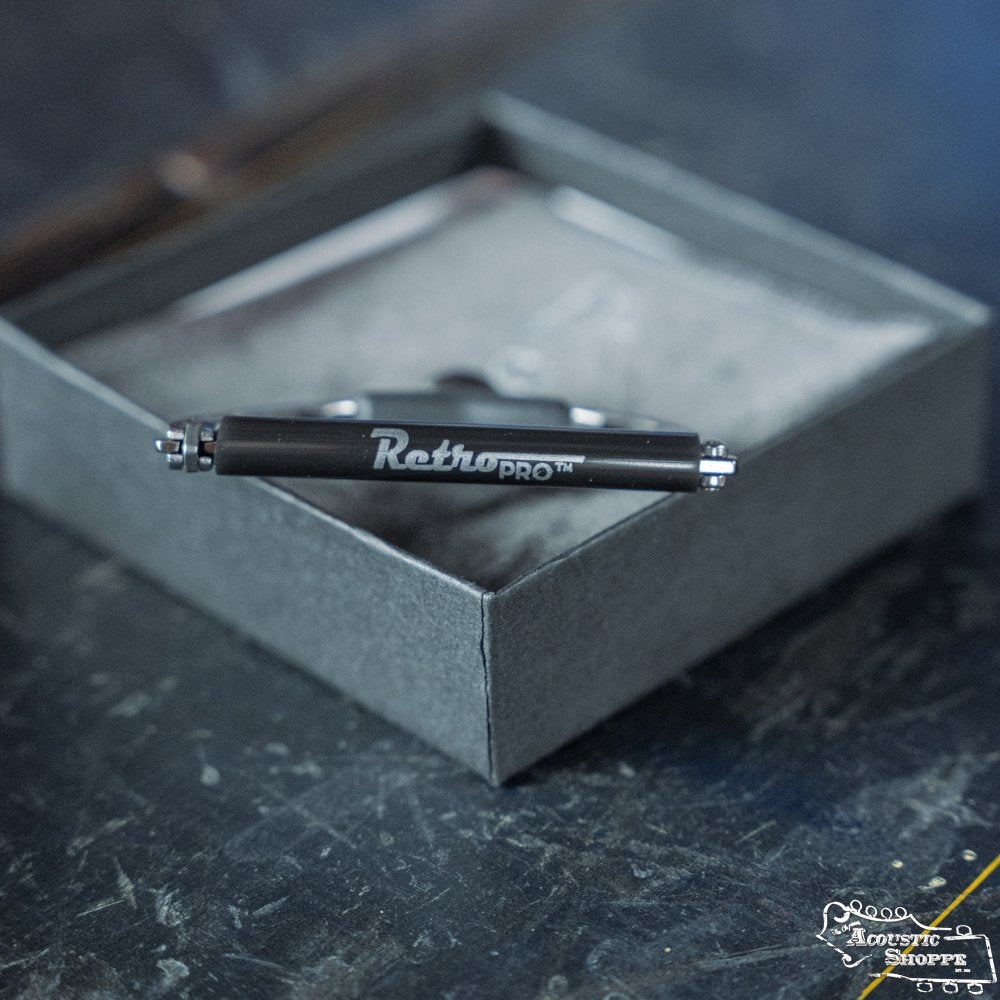 A Paige RetroPro 6-String Capo (PP-6-Retro) from Paige Musical Products is displayed atop an open gray box on a dark, textured surface, angled to showcase the brand name for a striking, modern look.