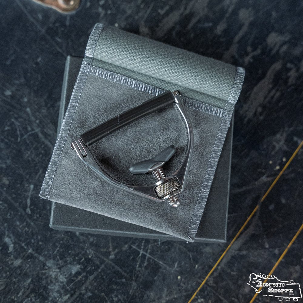 A Paige RetroPro 6-String Capo (PP-6-Retro) by Paige Musical Products rests on a folded gray pouch atop a dark, textured surface, with part of a logo visible in the bottom right corner.