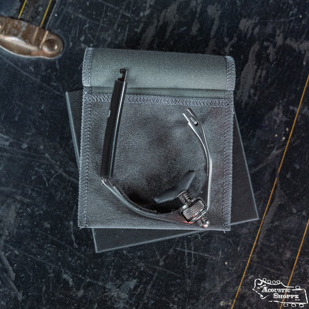 A black wristwatch with a metal strap rests partly unfastened on a pouch atop a box next to a Paige Musical Products RetroPro 6-String Capo (PP-6-Retro). Part of a boot appears in the corner, all on a scratched dark surface.