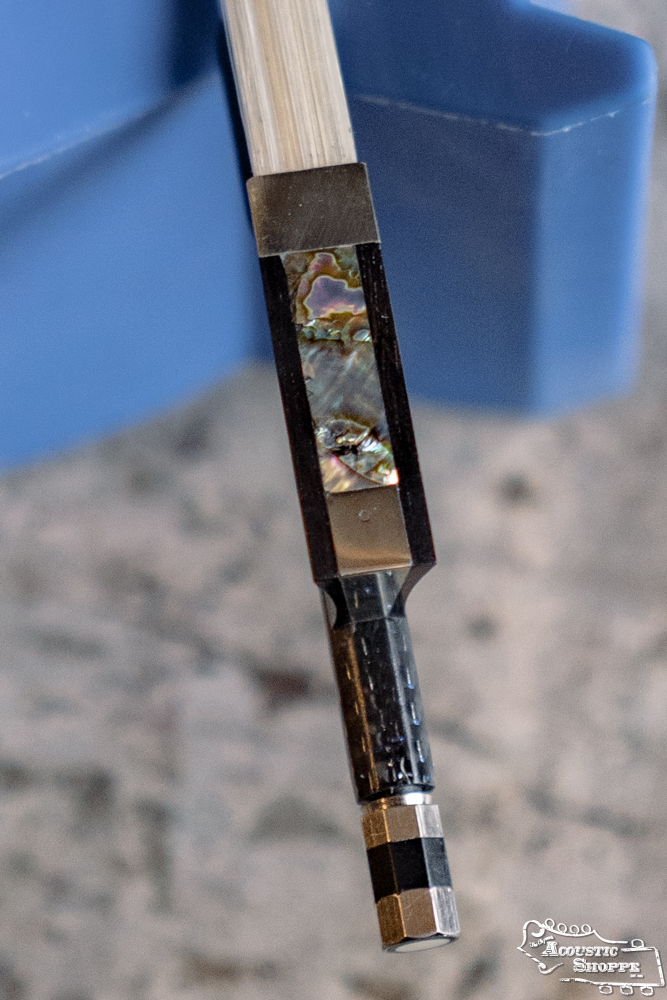 Close-up of the Howard Core Carbon 300 4/4 Violin Bow BLU's ebony frog with abalone inlay and silver fittings, resting near a blue, speckled object.