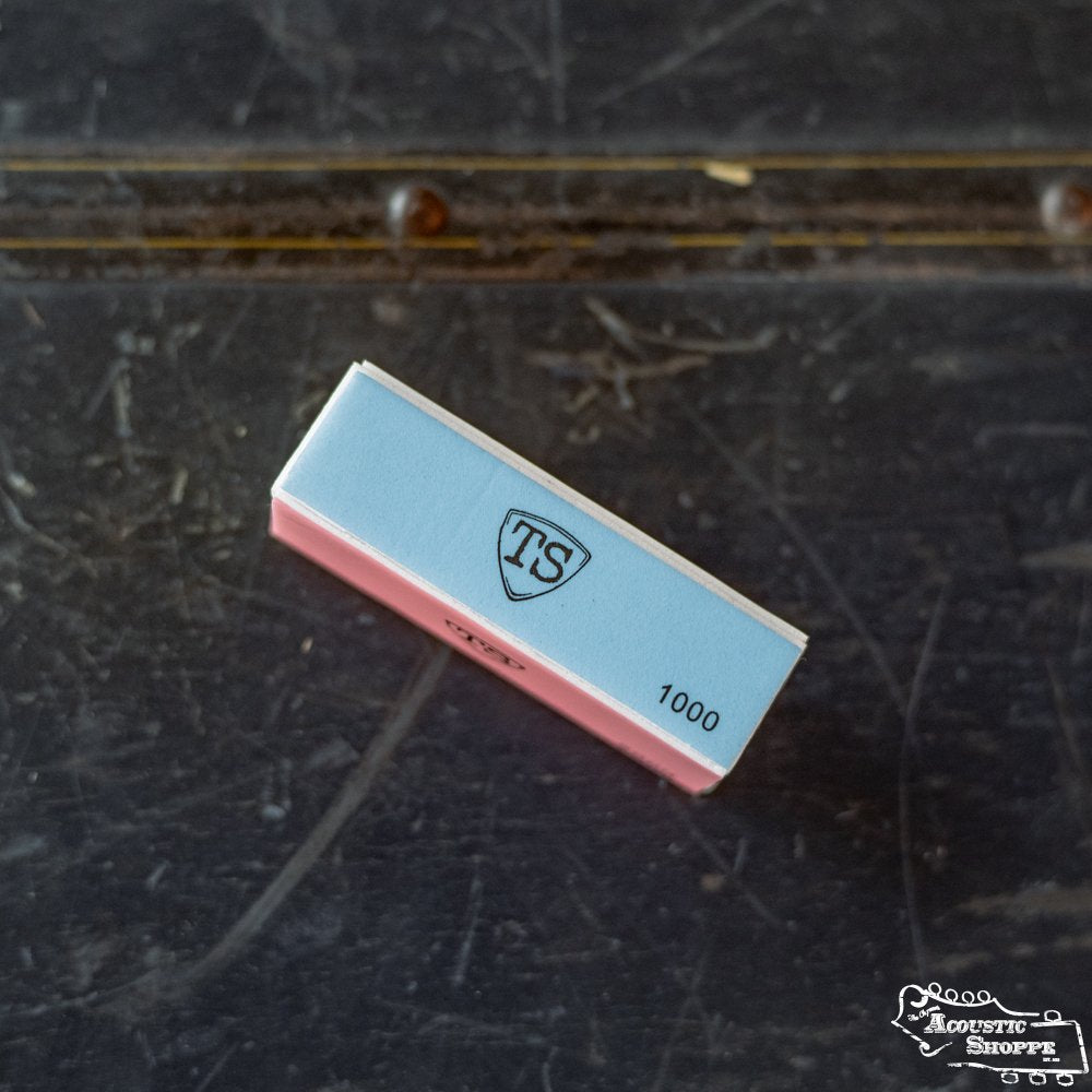 A small rectangular box with a blue and pink design, labeled ToneSlabs Pick Buffer and 1000, lies on a scratched dark surface—ideal for any acoustic shop. The ToneSlabs logo appears in the lower right corner.