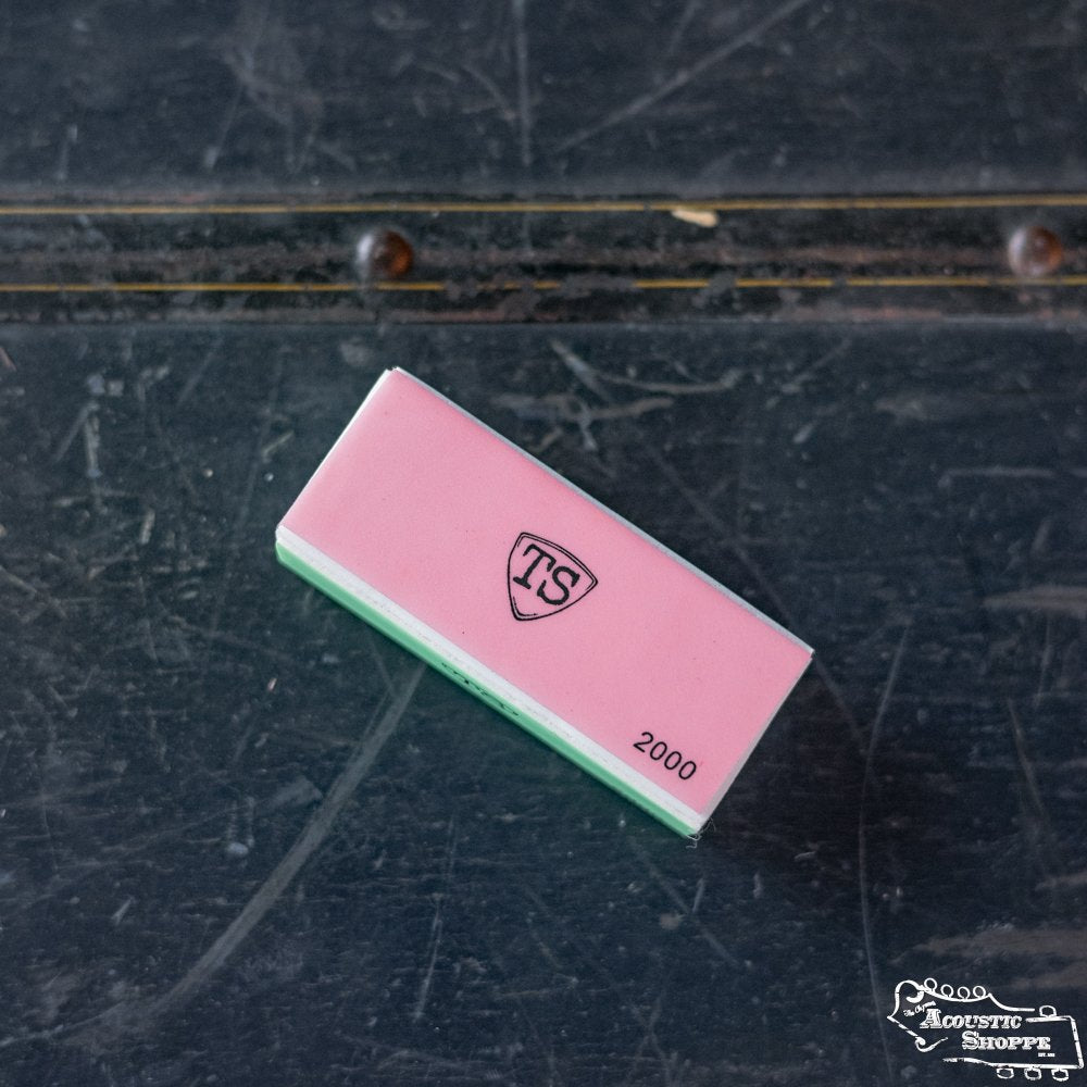 A rectangular pink and green ToneSlabs Pick Buffer sits on a worn dark surface with scratches, a brass strip, and signs of bevel polishing—possibly a tool found in an acoustic shop or used by an Eastman dealer.