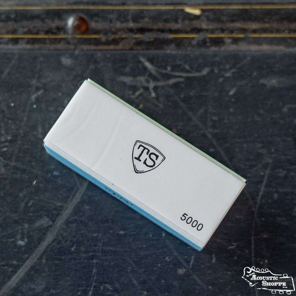 A rectangular ToneSlabs Pick Buffer box with a blue lower half and a white label featuring a shield-shaped TS logo and “5000,” resting on a dark, slightly worn surface—an ideal addition for any acoustic shop.
