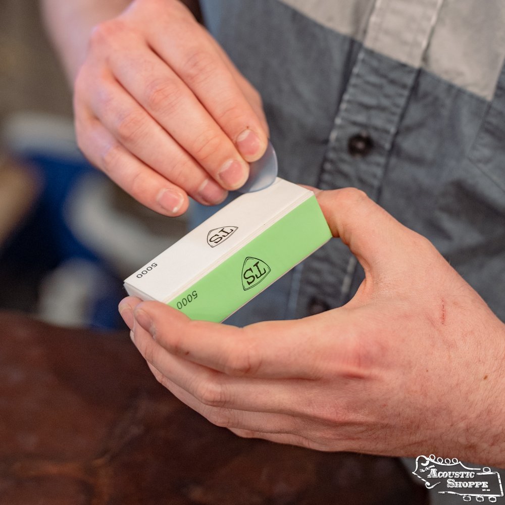 A person peels a sticker off a small, rectangular ToneSlabs Pick Buffer box with green and white packaging marked STS 5000, possibly from an Eastman dealer, while standing over a brown surface.