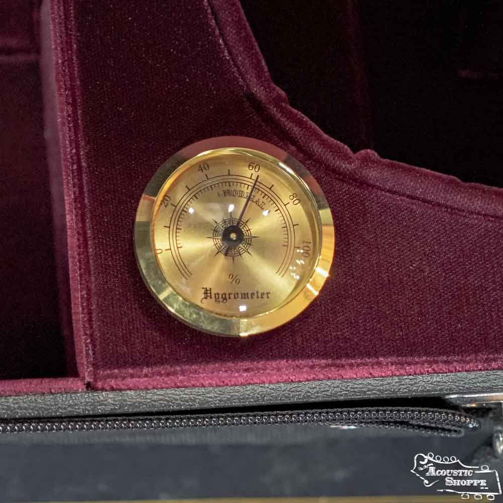 A round, gold analog hygrometer sits inside a Bobelock Velvet Suspension Half-Moon Violin Case (B1047VS-1). The burgundy velvet lining, a zipper, and part of the Bobelock logo are visible in the background.