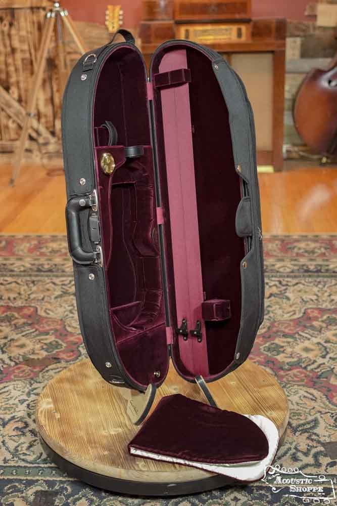 The Bobelock Velvet Suspension Half-Moon Violin Case B1047VS-1 by Bobelock is displayed open on a round wooden platform, showing its burgundy velvet interior, pockets, and compartments, with a matching accessory pouch in front. Musical instruments are in the background.