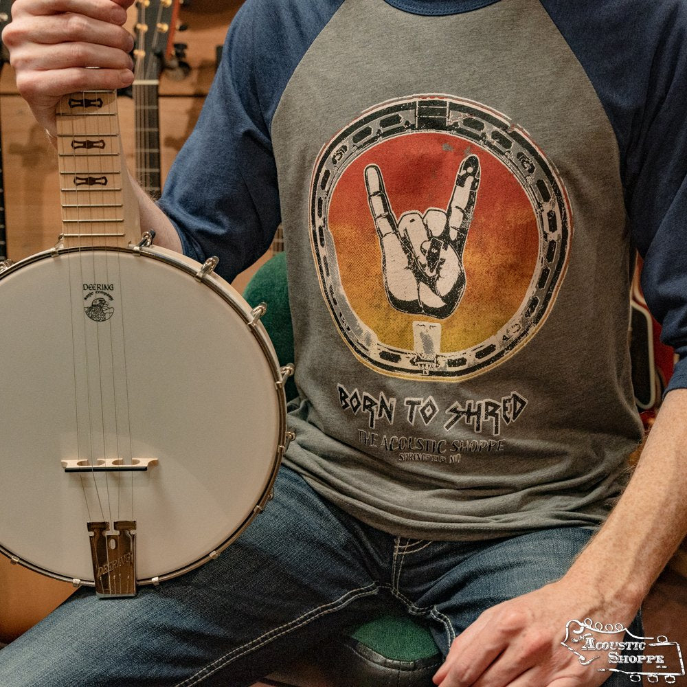 Someone in an Amplify Screen Printing & Promotions TAS Banjo Shred 3/4" Sleeve Baseball T-Shirt, featuring a hand making the rock sign in a circle, holds a white banjo with several guitars in the background.