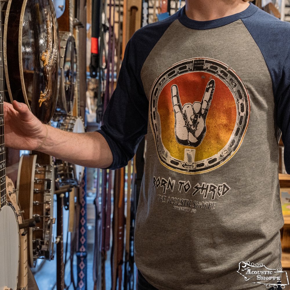 Wearing an Amplify Screen Printing & Promotions TAS Banjo Shred 3/4" Sleeve Baseball T-Shirt with a rock hand graphic, a person stands among hanging banjos and guitar straps in a music store.