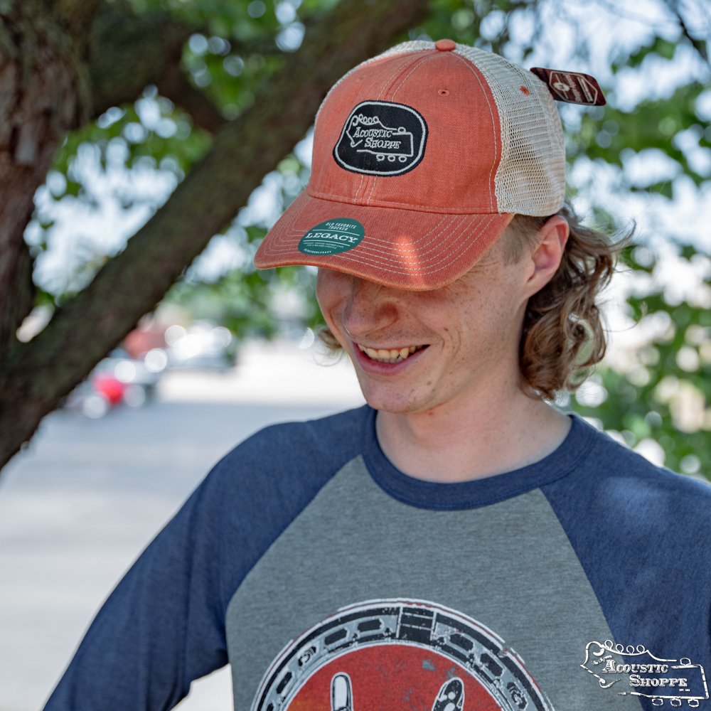 A young person with wavy blond hair smiles while wearing an Acoustic Shoppe Legacy Badge Trucker Cap in orange and black by Amplify Screen Printing & Promotions. Tree branches and blurred outdoor scenery appear in the background.