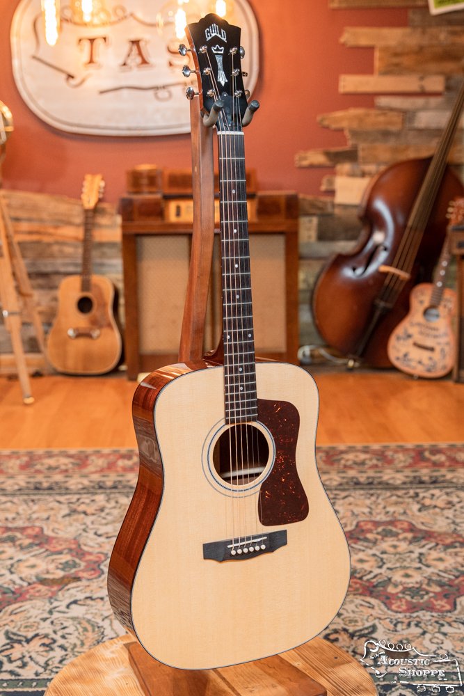 A Guild USA D-40 Traditional Sitka Spruce/African Mahogany Natural Top Dreadnought #1106 by Guild Guitars stands on a wooden stand in a cozy, warmly lit room with wood floors, a patterned rug, instruments, and rustic decor in the background.