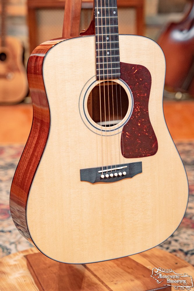 A close-up of the Guild USA D-40 Traditional Sitka Spruce/African Mahogany Natural Top Dreadnought #1106 by Guild Guitars, featuring a solid Sitka spruce top, dark pickguard, and six strings on a wooden stand over a patterned rug.