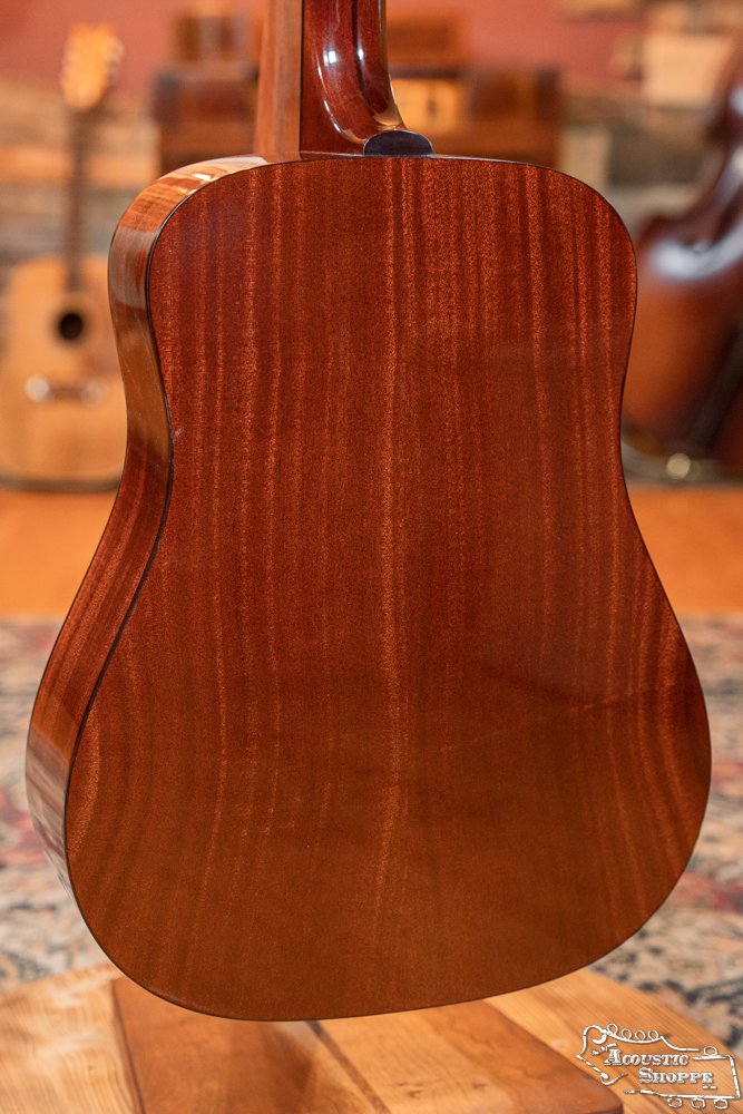 A close-up of the Guild USA D-40 Traditional Sitka Spruce/African Mahogany #1106 by Guild Guitars highlights its polished mahogany back and rich grain, with softly lit guitars and a warm room blurred in the background.