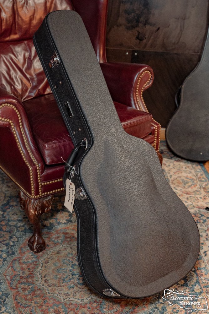 A Guild USA D-40 Traditional dreadnought guitar case with a handle and metal clasps stands upright against a red leather armchair with brass studs, on a patterned rug in warm light.