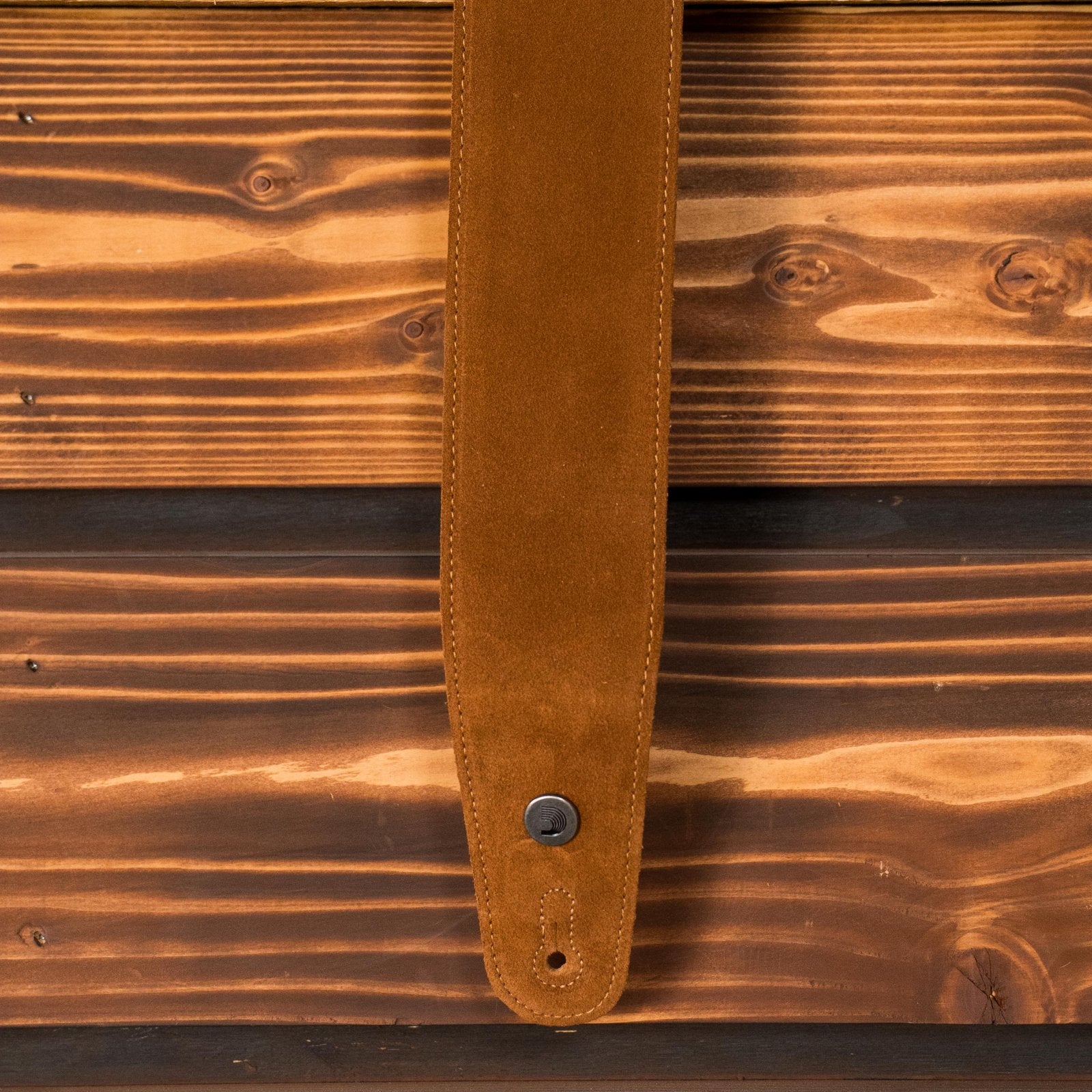 A close-up of the D'Addario 25SS02-DX Suede 2 1/5" Guitar Strap in Honey hanging vertically against a wooden wall, highlighting its comfortable design and the brand’s quality craftsmanship.